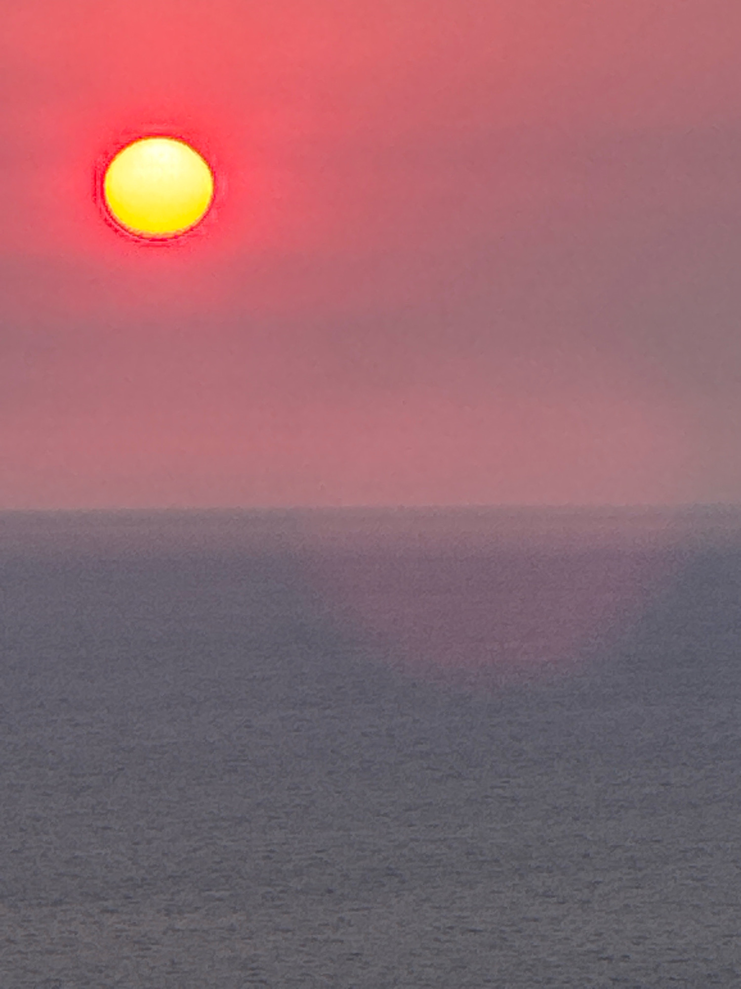A bright yellow sun with a red halo is visible in the upper left corner of the frame. The sun is positioned above a hazy horizon line. Below the horizon, the image depicts a body of water, likely the sea, with a textured surface. The sky is a gradient of pink and red hues, suggesting either a sunrise or sunset. The overall impression is one of a natural landscape, devoid of any man-made structures or human presence. The provided location of Zagora, Montenegro, is not discernible from the visual information within the image itself.
