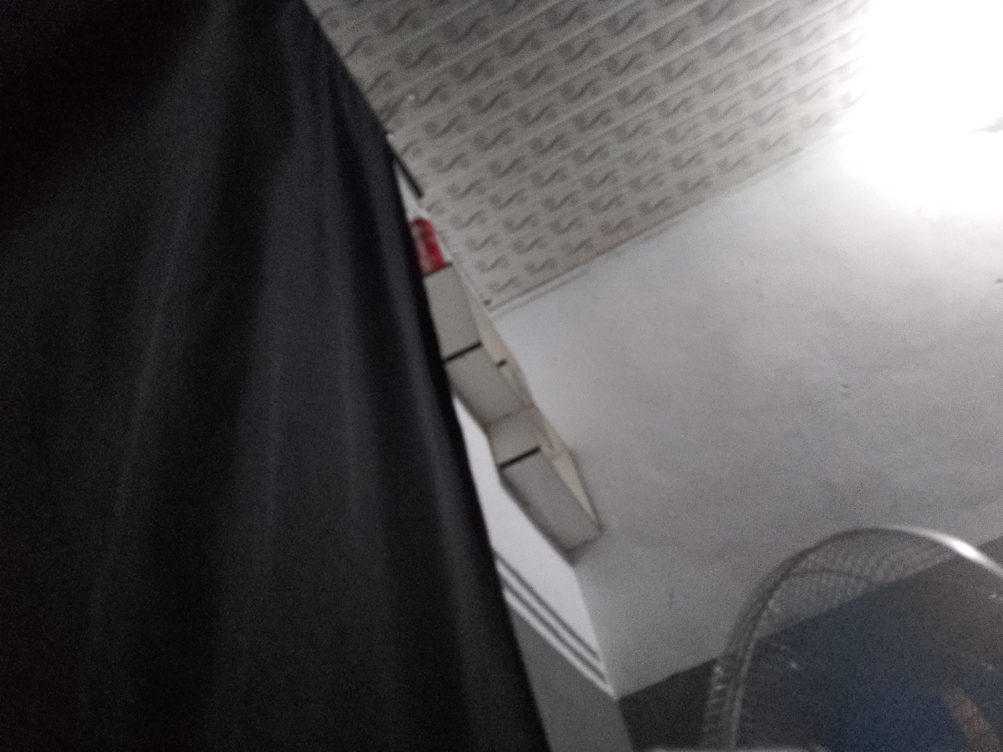 The image shows a close-up view of a black curtain or fabric on the left side, partially obscuring the background. The background consists of a white ceiling with a textured, patterned surface, possibly acoustic tiles, running horizontally across the top of the frame. Below the ceiling, a white wall is visible. In the lower right corner, the edge of a black fan with a metal grille is present. A sliver of a red object is visible behind the black fabric, near the ceiling. The lighting suggests an indoor environment with a light source illuminating the upper right portion of the scene. The image quality is grainy. This scene is not in Senti, Nigeria.