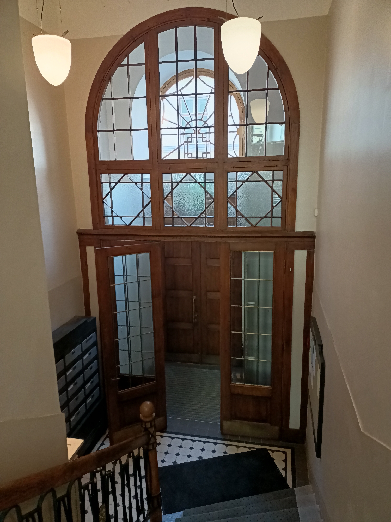 This is a high-angle, eye-level, indoor shot of a staircase and an entrance area, likely in an apartment building. The environment is characterized by wooden elements, including a large, arched window with a wooden frame and gridwork, and wooden doors with glass panels. The floor is tiled in a black and white geometric pattern.
In the foreground, a wooden handrail with metal spindles lines the staircase, which is carpeted. To the left, a dark filing cabinet with multiple drawers is visible. The focal point of the image is the set of double wooden doors, one of which is ajar, revealing a hallway beyond. Above the doors, a substantial wooden-framed window is segmented into numerous panes, some of which are frosted or textured glass, and others are clear glass. Two pendant lights hang from the ceiling, one on the left and one reflected in the window. The walls are painted in a neutral, light color.
The scene appears to be located in Tallinn, Estonia, a city known for its historic architecture which often features similar wooden detailing and materials. The overall impression is one of an older, well-maintained building. No people are present in the image.