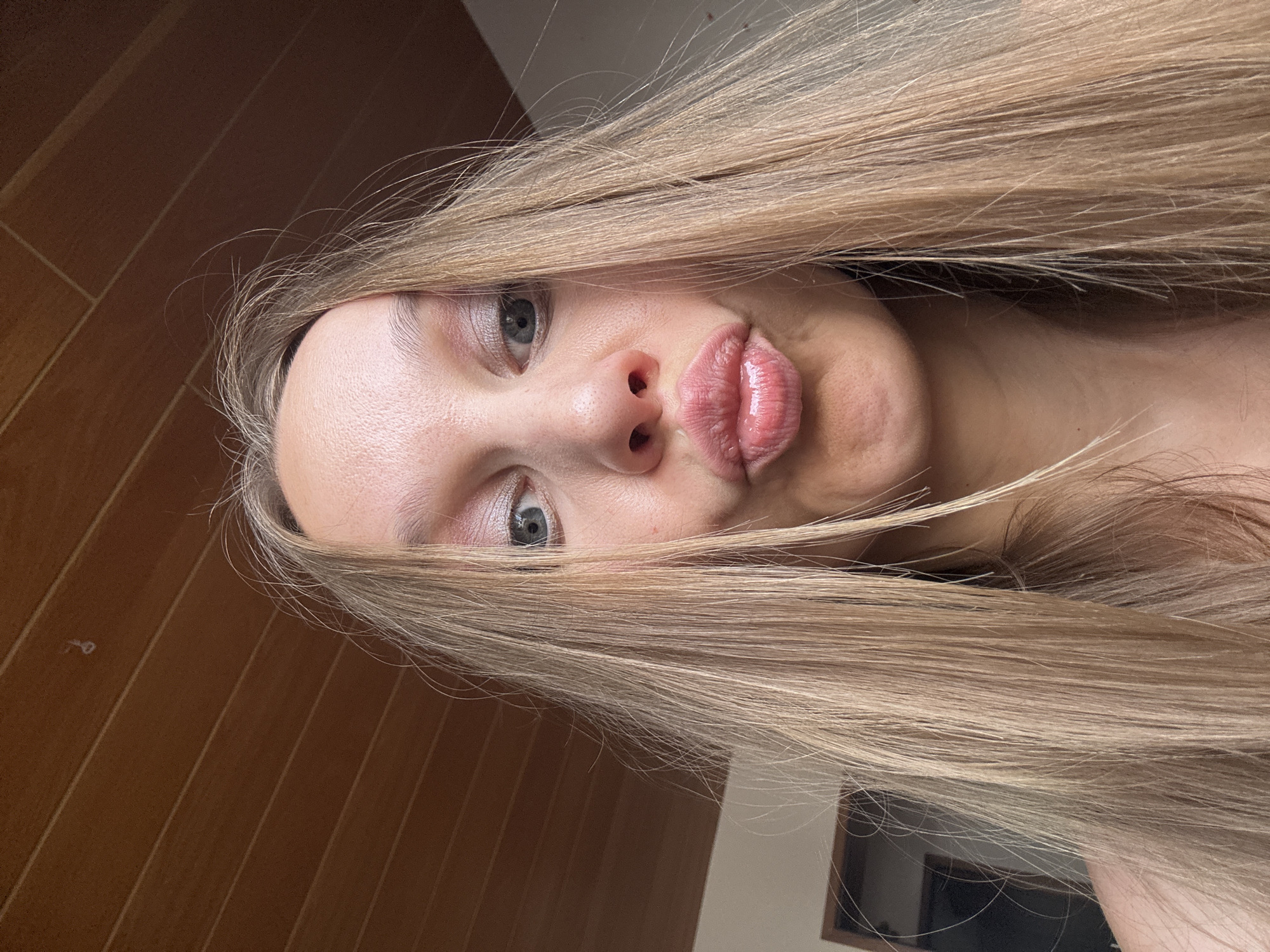 A young blonde woman with long, straight hair is posing for a selfie. She is wearing eye makeup and her lips are puckered. The background appears to be a wooden ceiling and a wall. The woman's face fills most of the frame, and her hair is falling around her face and shoulders. There is no indication of the specific location in the city of Überherrn, Germany.