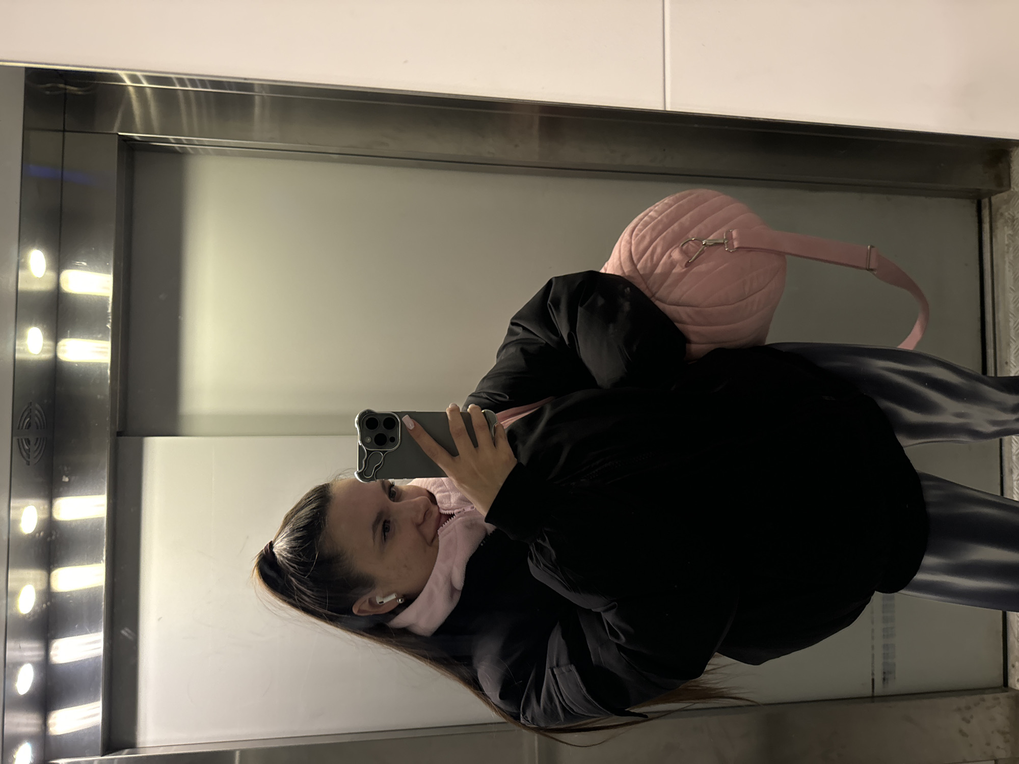 A woman is taking a selfie in what appears to be an elevator. She is wearing a black jacket over a pink garment, and a pink bag is slung over her shoulder. She is holding a grey smartphone to take the photo. The elevator's interior is illuminated by overhead lights. The location is implied to be in the city of Odintsovo, Russia, but this is not directly evident from the image itself.