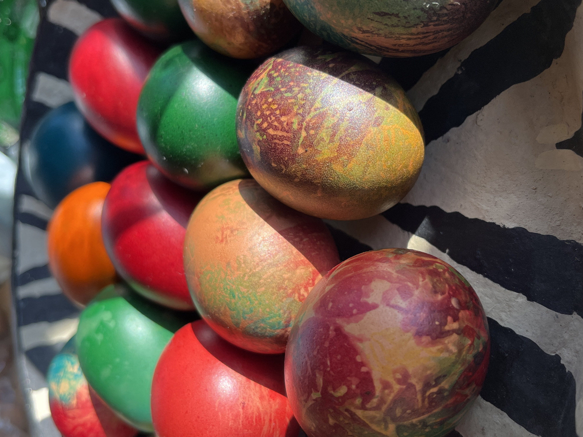 The image depicts a close-up view of a stack of colorful wooden spheres, possibly decorative ornaments. The spheres are arranged in rows and display a range of solid colors, including red, green, orange, and blue, along with marbled and patterned surfaces that blend multiple hues. The spheres rest on a surface with a black and white striped pattern reminiscent of zebra stripes. The lighting suggests an outdoor setting, with direct sunlight highlighting some spheres and casting shadows on others. The location is identified as Pchelnik, Bulgaria. There is no sign of any people in this picture.