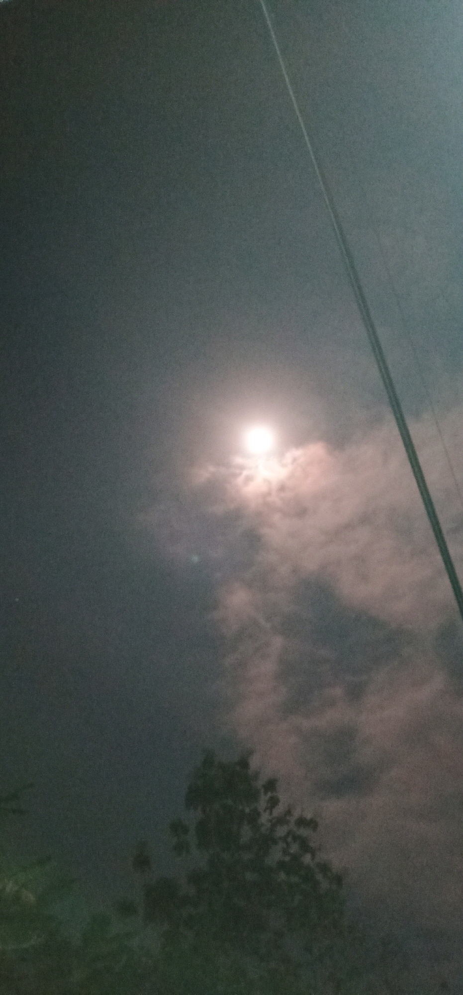 The image captures a nighttime scene in Gambarsari, Indonesia, featuring a bright, full moon partially obscured by clouds. The moon is the dominant light source, casting a soft glow on the surrounding sky and clouds, which appear wispy and textured. In the foreground, the silhouette of trees is visible, suggesting a natural or semi-natural environment. A utility pole with wires extends diagonally across the right side of the frame, indicating the presence of infrastructure within the urban or suburban setting. The overall atmosphere is serene and slightly mysterious due to the low-light conditions and the obscured moon.
