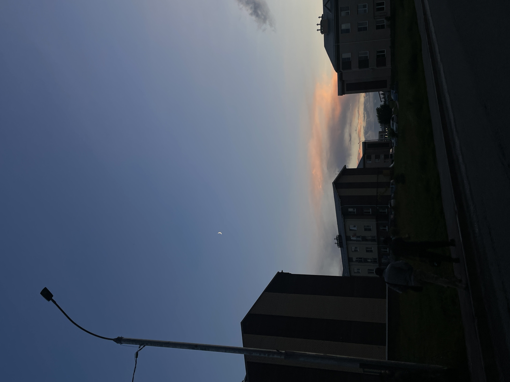 The image depicts a street scene in the city of Zverevo, Russia, during twilight. A tall, curved lamppost is visible on the left side of the frame. In the background, several apartment buildings are silhouetted against a dusky sky. The sky features a faint crescent moon and streaks of soft pink and orange clouds from the setting sun. Two figures, appearing as dark silhouettes, are walking away from the camera on a sidewalk in front of the buildings. The foreground shows a paved road and a grassy area. The overall atmosphere is calm and subdued, characteristic of early evening in an urban environment.