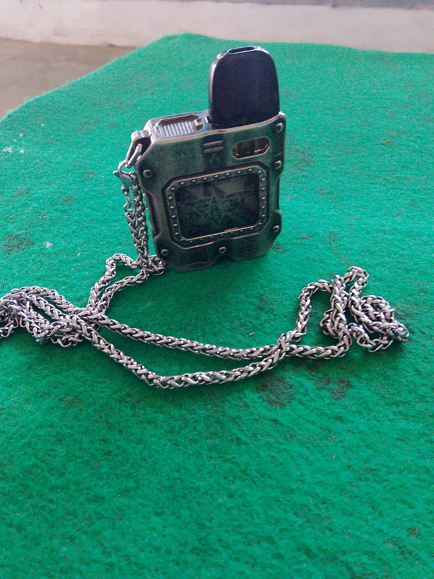 A close-up, high-angle shot shows a metallic vape device with a dark mouthpiece and a chain necklace lying on a green, fuzzy surface, possibly a rug or felt. The vape device has a rectangular screen and a rugged, industrial design, with visible screws and textured metal plating. The chain is thick and silver-colored, with a woven or braided appearance, draped around the vape device and extending towards the bottom of the frame. The background is blurred, showing a light-colored surface and the edge of the green surface. The lighting appears to be natural, casting soft shadows. The scene is static and does not depict any action or interaction. The location is not specified in the image or its context.