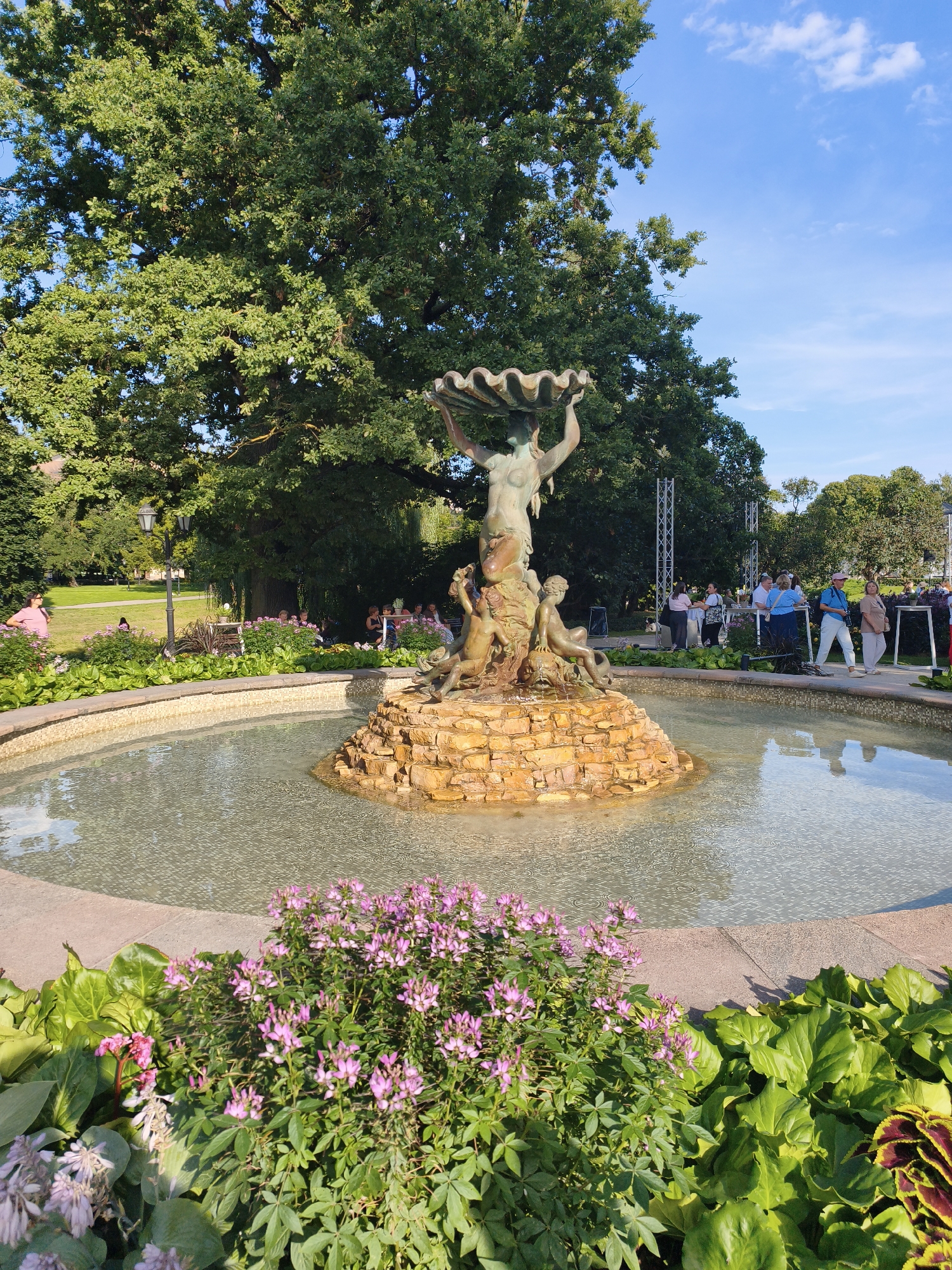 The image depicts a bronze fountain sculpture in a circular pool, surrounded by lush green foliage and pink flowers. The sculpture features a central figure holding a large shell above its head, with smaller figures at its base. The pool has a textured bottom and is bordered by stone. In the background, several people are visible in a park-like setting, with trees and a bright blue sky dotted with white clouds. The scene appears to be a public gathering or event. The fountain and surrounding park are located in Vērmanes Garden, a historic public park in Riga, Latvia.