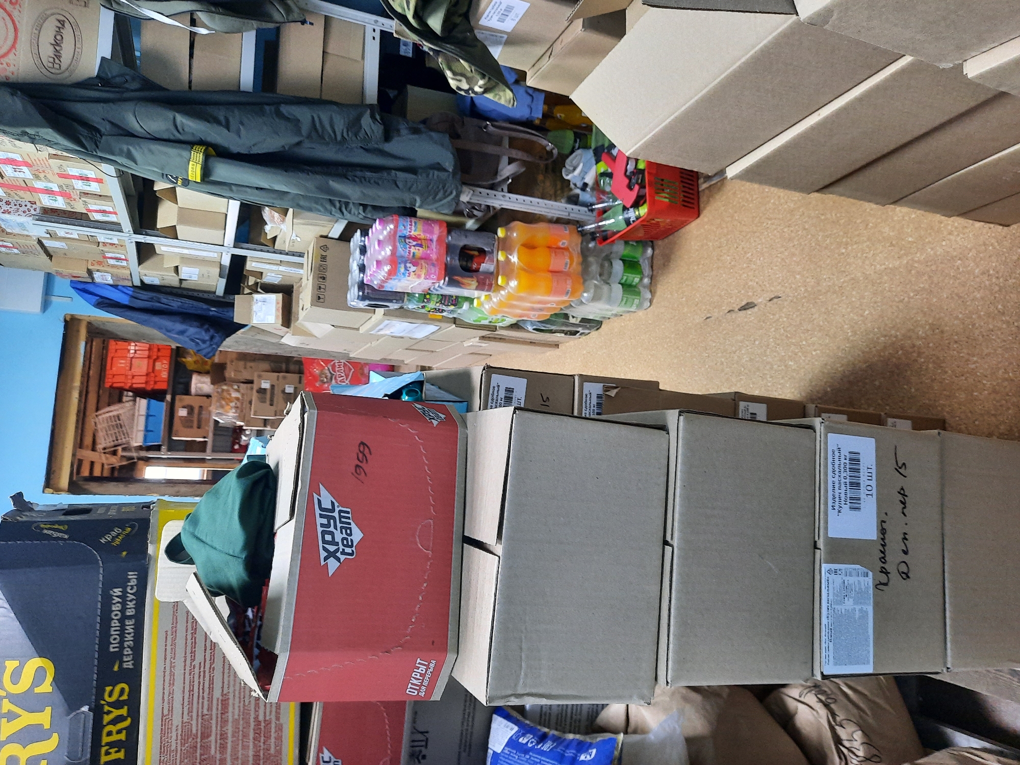 The image shows a storage area, likely in Krasnogorskoye, Russia, filled with various items. There are numerous cardboard boxes stacked in rows. Some boxes have labels and writings on them, including brand names like "Fry's," "Хрус Team", and "Вакконд." The text on some boxes is in Russian. Some boxes contain bottles of drinks, while others appear to hold other items. Some items on the shelves include clothing. A red plastic crate can also be seen.