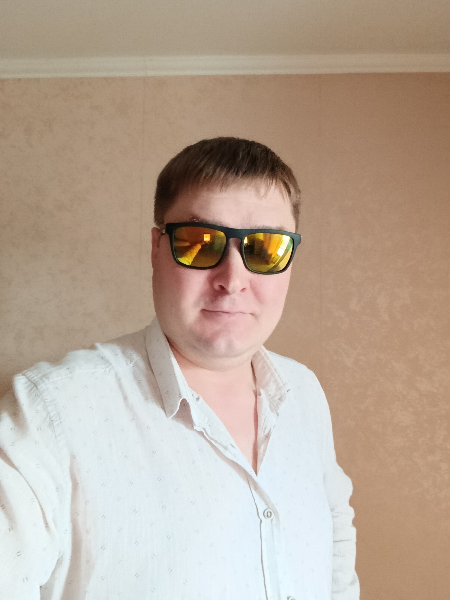 The image shows a medium shot of a man wearing sunglasses and a white button-down shirt. The man is positioned in the center of the frame, looking directly at the camera. He has short, light brown hair and is wearing black-framed sunglasses with orange-tinted lenses that reflect a bright light and possibly part of his surroundings. The shirt he is wearing is white with a subtle pattern of small, repeating diamond shapes. The background is a textured wall in a light brown or beige color. The lighting suggests an indoor setting, likely a room. The image's overall composition is a selfie-style portrait. The man appears to be standing in a room in Petropavl, Kazakhstan, though no specific landmarks are visible in the image.