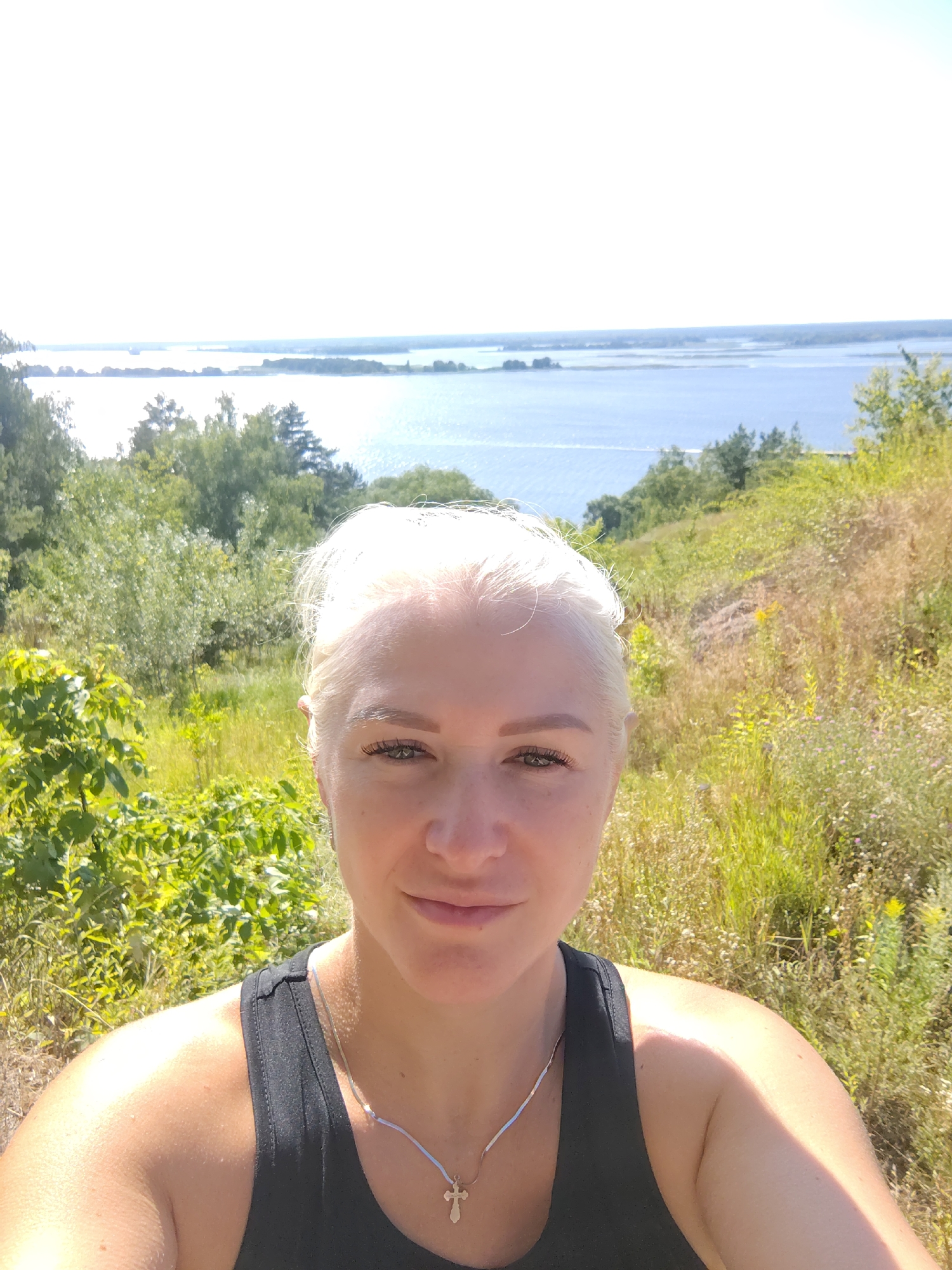 A selfie taken from a slightly low angle shows a woman with blonde hair and green eyes. She is wearing a black tank top and a silver necklace with a cross pendant. She is smiling and looking directly at the camera. The background features a body of water, likely a lake or river, with trees and vegetation on the slopes leading down to it. The sky is clear and bright, suggesting it is daytime. The location is identified as Staiky, Ukraine.
