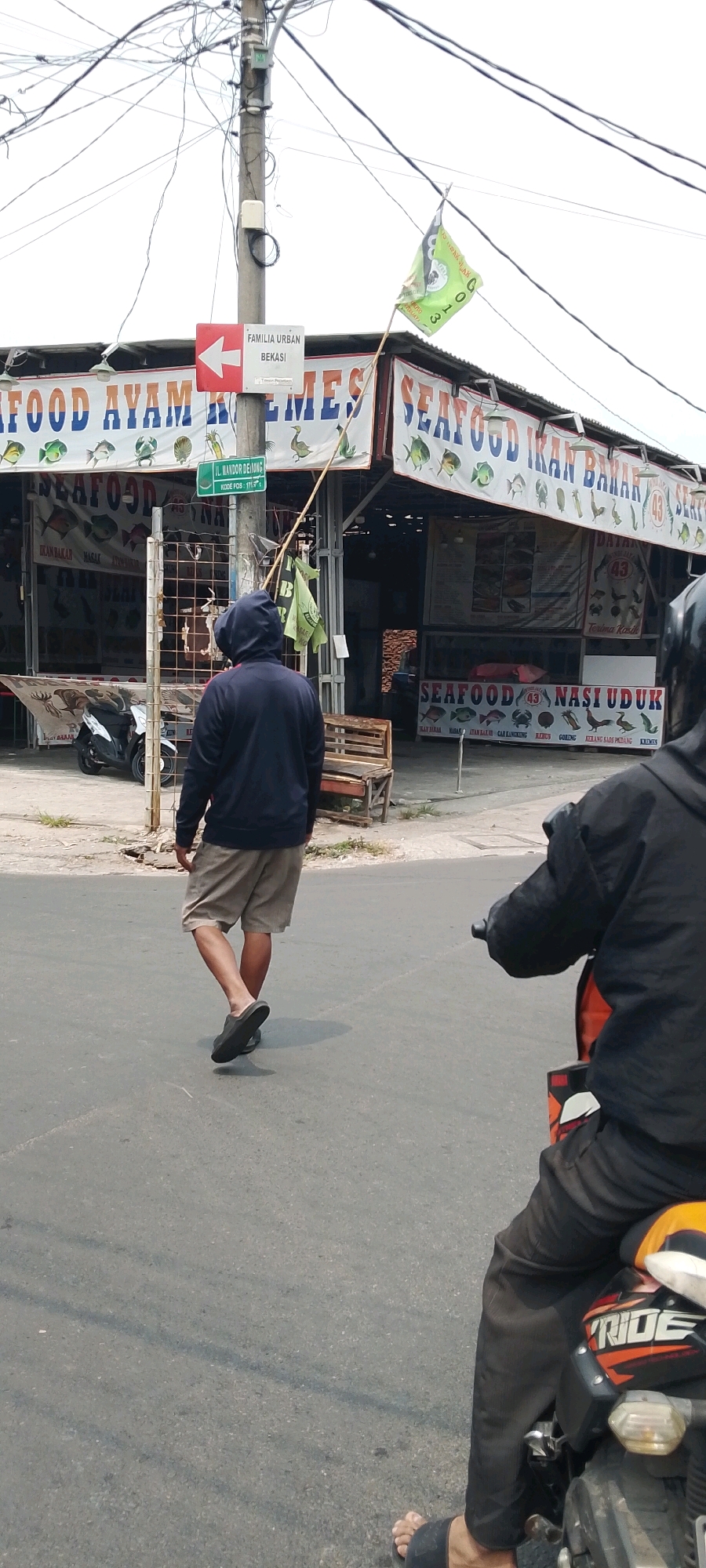 A medium shot captures a street scene in Bekasi, Indonesia. A person wearing a dark hooded jacket and beige shorts walks away from the camera, their back to the viewer. They are wearing dark sandals. To the left, a scooter is parked. A lamppost with overhead wires stands prominently in the foreground. A sign on the lamppost reads "FAMILIA URBAN BEKASI" with an arrow pointing left. A green flag is attached to the lamppost. In the background, a building with banners displaying "SEAFOOD AYAM KEMES" and "SEAFOOD IKAN BAHAR" is visible, indicating a food establishment. Another person is partially visible on a motorcycle in the lower right corner of the frame. The street is paved, and the sky is overcast.
