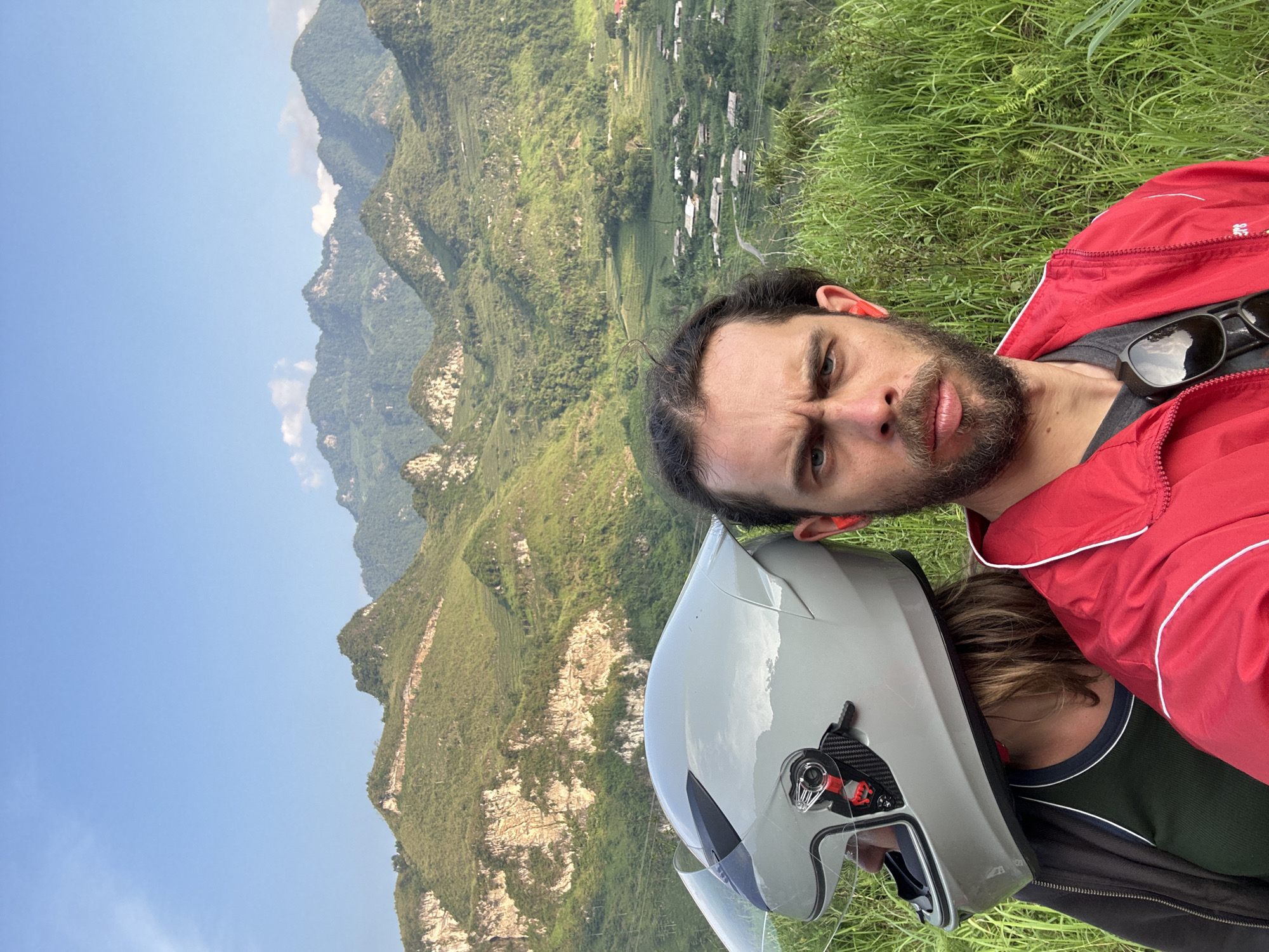 The image captures a man and a child with a motorcycle helmet outdoors in Du Già Commune, Vietnam. The man, likely in his 30s, has dark hair, a beard, and a red jacket. He is positioned in the foreground, looking at the camera with a slightly furrowed brow. Beside him, a child wears a motorcycle helmet.
The background reveals a mountainous landscape with lush green vegetation, and a settlement can be seen in the distance. The sky is clear and blue. The presence of grass in the foreground suggests they are standing on a hillside.