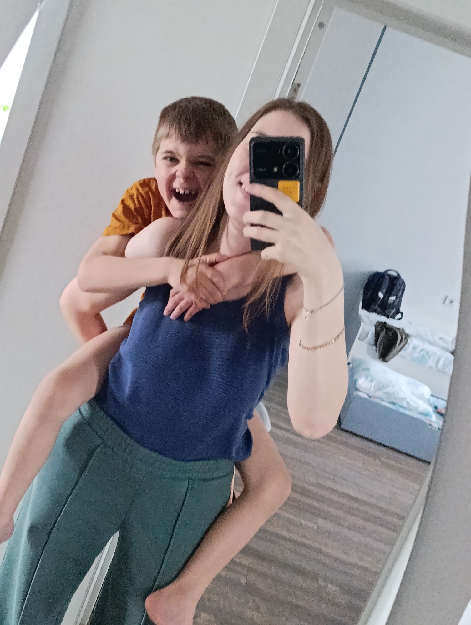 This mirror selfie captures a joyful moment between a woman and a young boy, likely within a home in Chaikovsky, Russia. The woman, with long brown hair, is wearing a blue tank top and green sweatpants. She holds a black smartphone with a distinctive yellow section on its back, taking the photo in front of a mirror. Her face is partially obscured by the phone, but her wide smile and visible eyes convey happiness.
A young boy, with short brown hair and an orange t-shirt, is riding piggyback on the woman, his arms wrapped around her neck. He is laughing heartily with his mouth open, showcasing a playful and exuberant expression. Both subjects appear to be in high spirits, enjoying a casual and fun interaction.
The setting visible in the mirror's reflection suggests an indoor environment with light-colored walls and a light brown wooden floor. In the background of the reflection, a bed with light-colored bedding is visible, along with a black backpack and possibly other personal items on or near the bed. The time of day is not explicitly clear, but the interior lighting suggests a well-lit space. There is no discernible text visible in the image.