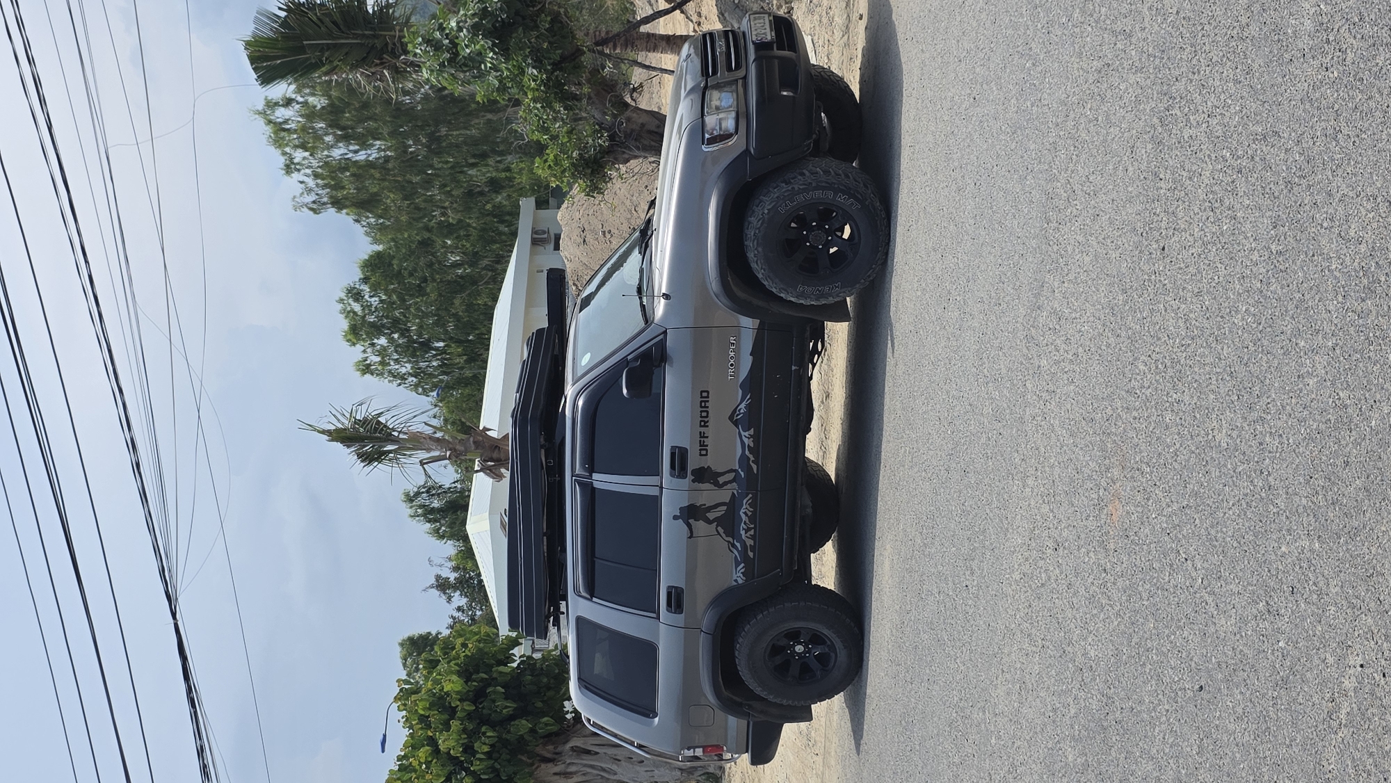 The image, taken in Bac Nha Trang Ward, Vietnam, displays a grey Isuzu Trooper SUV parked on the side of an asphalt road under bright daylight. The vehicle is heavily modified for off-road adventures, featuring prominent black alloy wheels fitted with Kenda Klever M/T tires. A robust black roof rack system supports two large black storage boxes or a rooftop tent on the vehicle's roof.
The side of the SUV is adorned with custom decals, including the bold text "OFF ROAD," below which are silhouettes of jagged mountains and two figures appearing to be hikers or adventurers. The model name "TROOPER" is also visible on the side door. The front license plate, "80A-023.30," is partially visible.
The surrounding environment suggests a warm climate, with lush green trees and several palm trees lining the road. A light-colored building is partially visible in the background. Numerous black power lines crisscross the clear to partly cloudy sky above. The asphalt road surface appears well-maintained. There are no people visible in the shot, and the vehicle appears stationary.