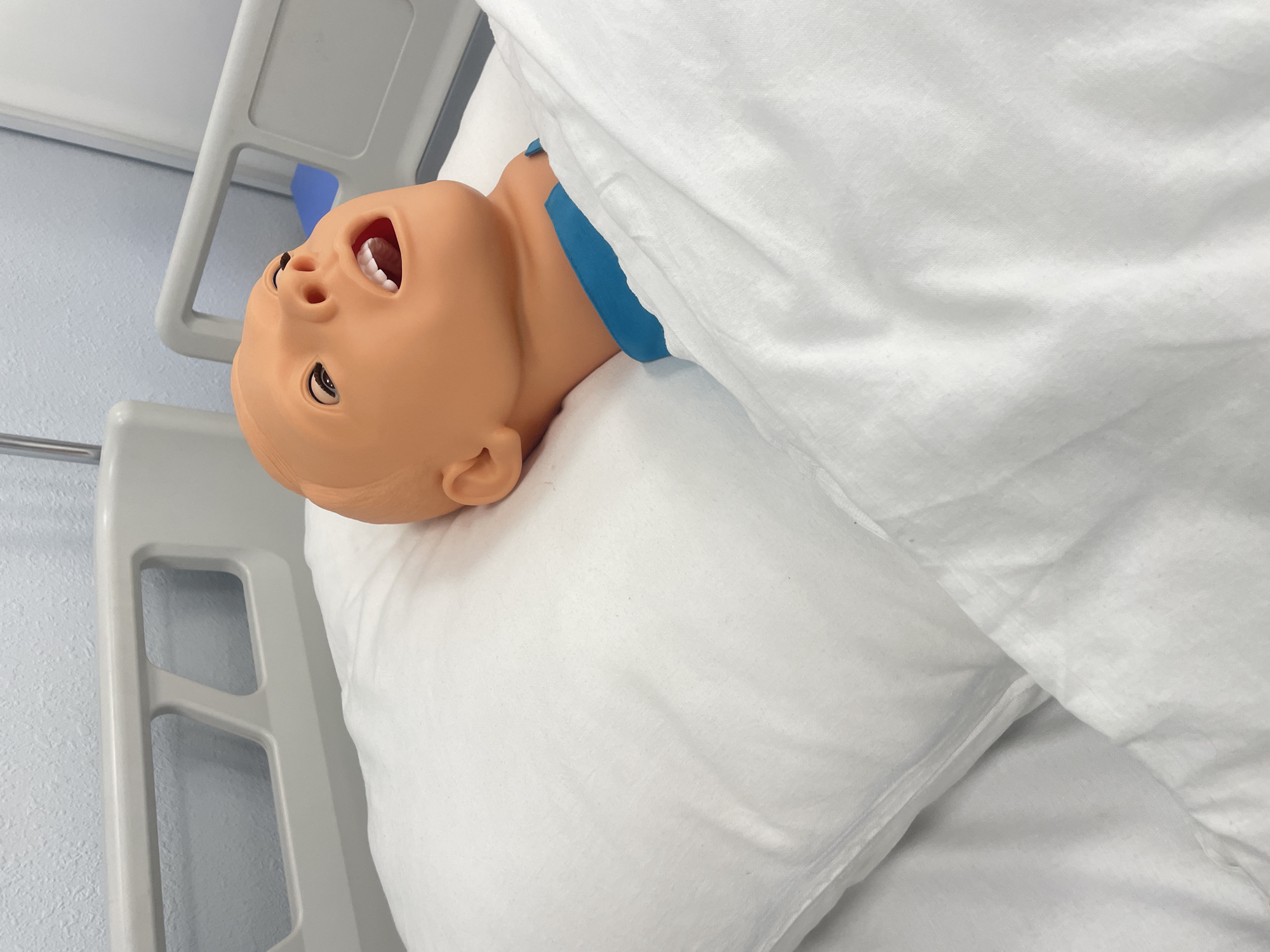 A light-skinned infant mannequin with a blue band around its neck lies in a hospital bed. The mannequin's mouth is open. The mannequin is covered with a white blanket and rests on a white pillow. The hospital bed has white railing. The wall behind the bed is light blue. The location and city are unknown.