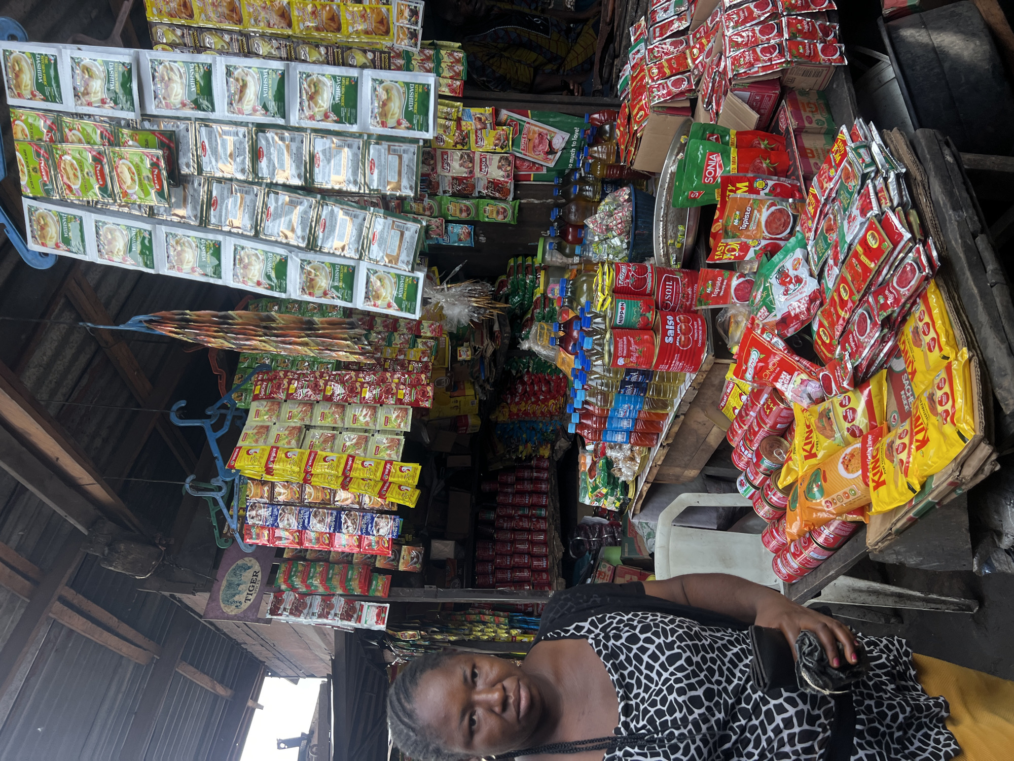 A medium shot captures a woman standing in front of a market stall in Nkpor, Nigeria. The stall is laden with various food products, predominantly packaged seasoning powders, tomato paste sachets, and canned goods. The woman, with dark skin and braided hair, wears a black and white patterned top and holds a black purse in her left hand. She looks directly at the camera with a neutral expression. Behind her, the stall is densely packed with goods arranged on shelves and hanging displays. Rows of red-colored cans are visible, along with numerous colorful packets of spices and sauces. Bottles of liquid, likely cooking oil or beverages, are also present on a lower shelf. The market appears to be an open-air setting with a corrugated metal roof supported by wooden beams. The lighting suggests it is daytime, with overcast conditions. The overall impression is a bustling market environment with a focus on retail of food products.