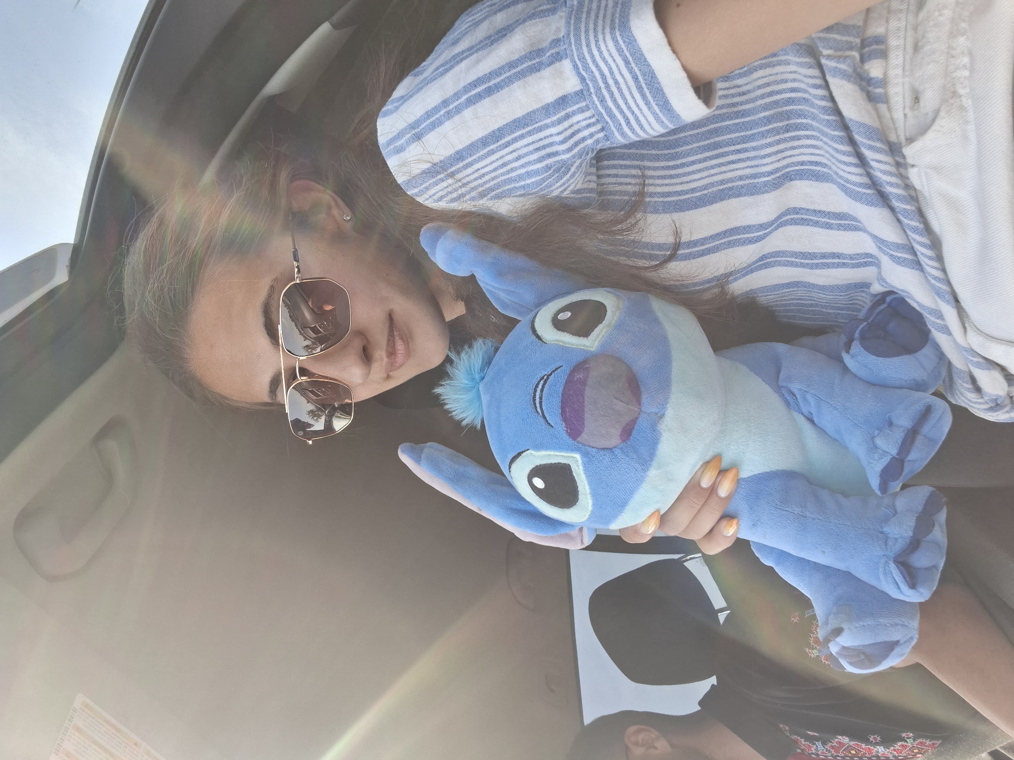 A young woman, wearing sunglasses and a blue and white striped shirt, sits in a car. She is holding a blue Stitch stuffed toy. The woman has long brown hair and her nails are painted with an ombre design of orange and yellow. A man is visible in the background, to the left of the woman. The car interior is dimly lit, with sunlight streaming in from the side. The background outside the car is blurred.