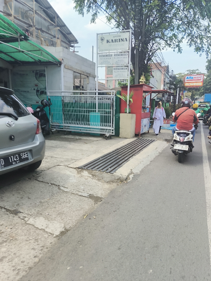 The image shows a street scene in Ngamprah, Indonesia, located at coordinates -6.861 latitude and 107.501 longitude. A silver Toyota Yaris is parked on the left side of the street, facing the camera. A person in an orange shirt is riding a motorbike on the right side of the street, while a person in white clothing is walking on the sidewalk on the right side. There is a red kiosk on the right side of the street with a sign above it that reads “Apotik Karina”. Behind the kiosk, there is a green fence with a green awning above it. There are trees and other buildings in the background. There is a metal drainage grate on the street between the parked car and the motorbike. The sidewalk is lined with a concrete curb and a row of drainage grates. The street is paved with asphalt and has a white painted line down the center.