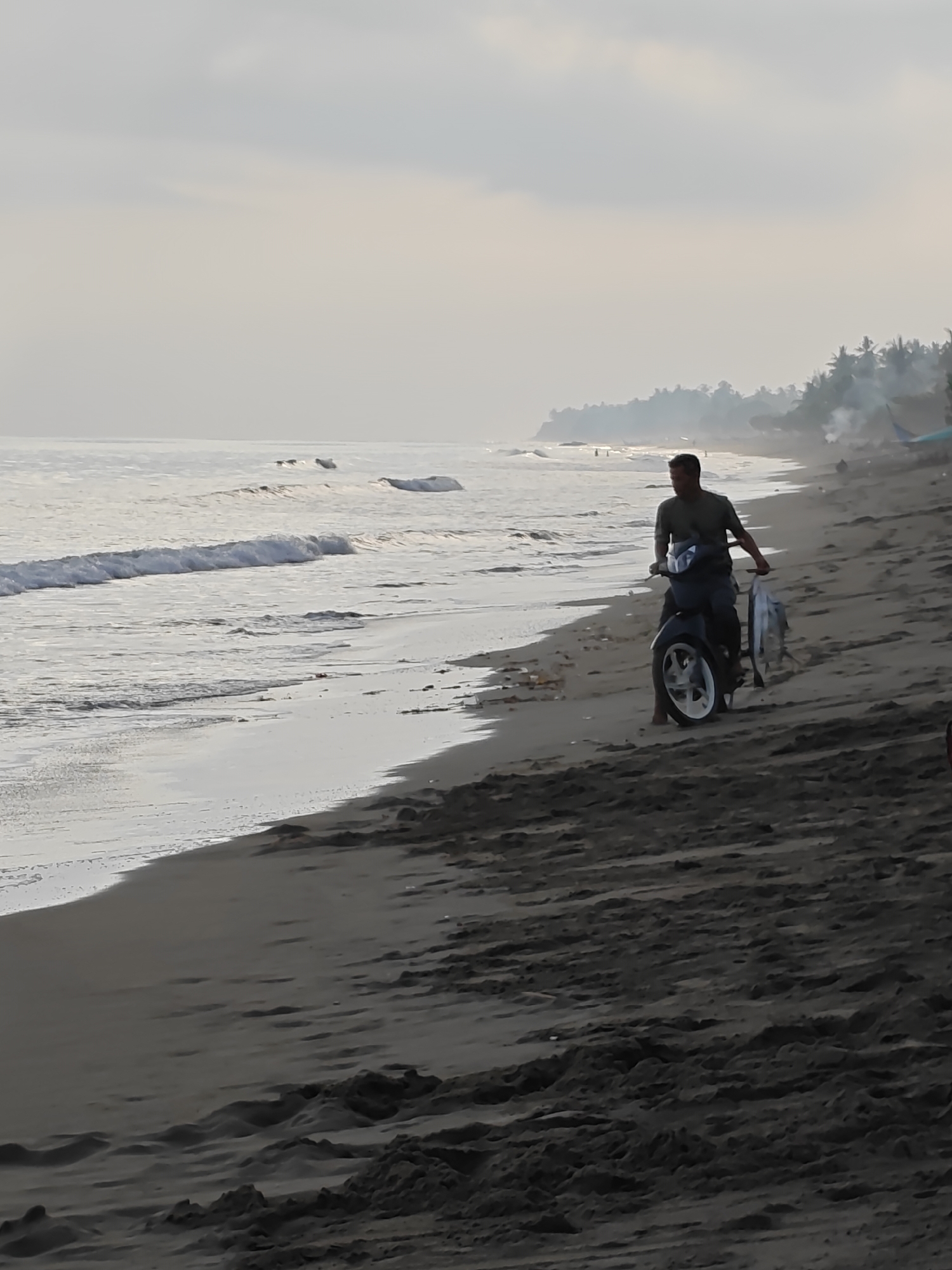 In Curehtunong, Indonesia, on a beach with dark sand, a man rides a dark-colored scooter. He is wearing a dark green t-shirt and dark pants. He is carrying two large fish, which are hanging from his right hand. The scooter has a white rim on the rear wheel. The ocean is to the left of the man, with gentle waves washing onto the shore. In the distance, palm trees line the coast. The sky is overcast with a pale, hazy light. There are some small pieces of debris on the sand near the water's edge.