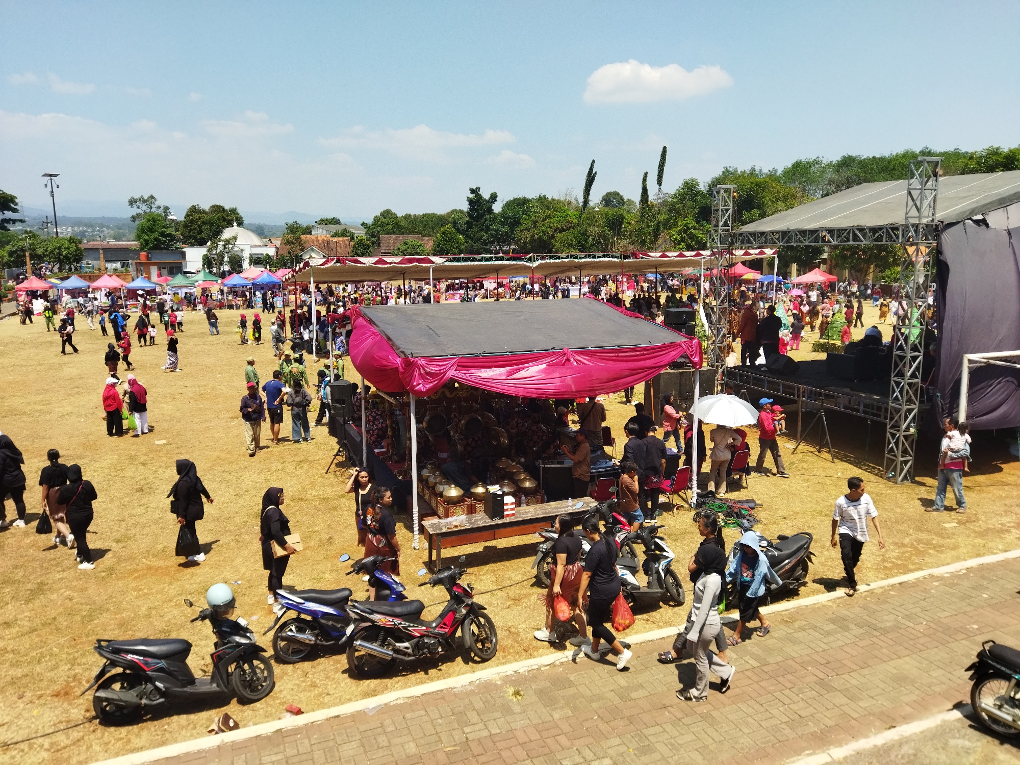 An outdoor daytime event is taking place on a large, dry, grassy field. Numerous people are milling about, interacting with various stalls and a stage. The sky is blue with scattered clouds. In the background, trees and buildings suggest a semi-urban or rural setting. A stage structure with metal scaffolding and a large dark tarp is visible on the right side of the frame. Several temporary stalls with colorful awnings are set up across the field, displaying goods and attracting visitors. Motorcycles are parked in various locations, indicating attendees arrived by vehicle. The overall atmosphere appears lively and social, with people engaged in diverse activities such as browsing, walking, and conversing.