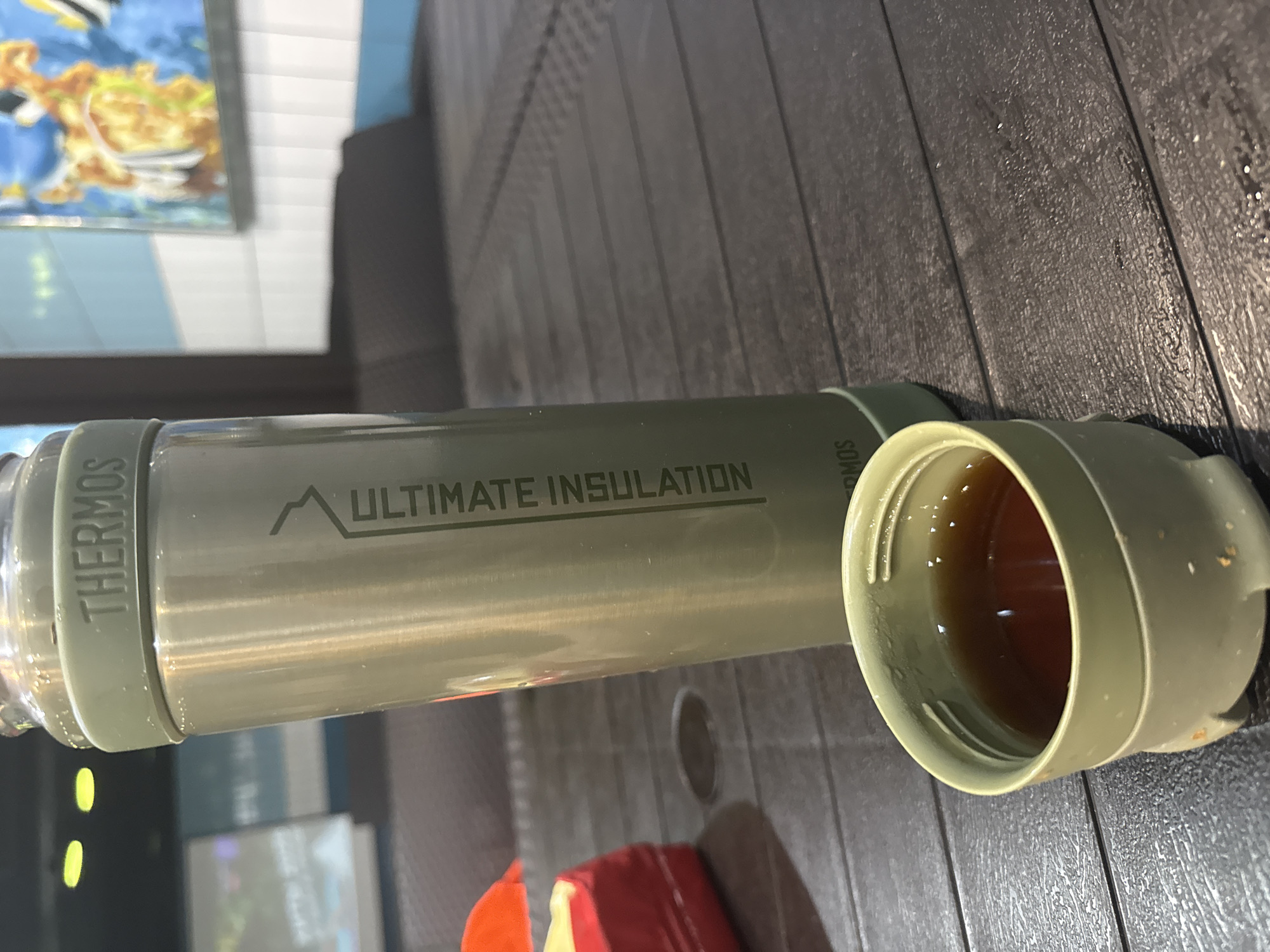 A green Thermos brand bottle labeled "ULTIMATE INSULATION" lies horizontally on a dark, textured surface. The bottle's lid is detached and positioned next to it, revealing a dark liquid inside. The background is blurred, suggesting an indoor setting with a painting and possibly a wall. The image does not contain enough information to determine the location is Tobolsk, Russia.