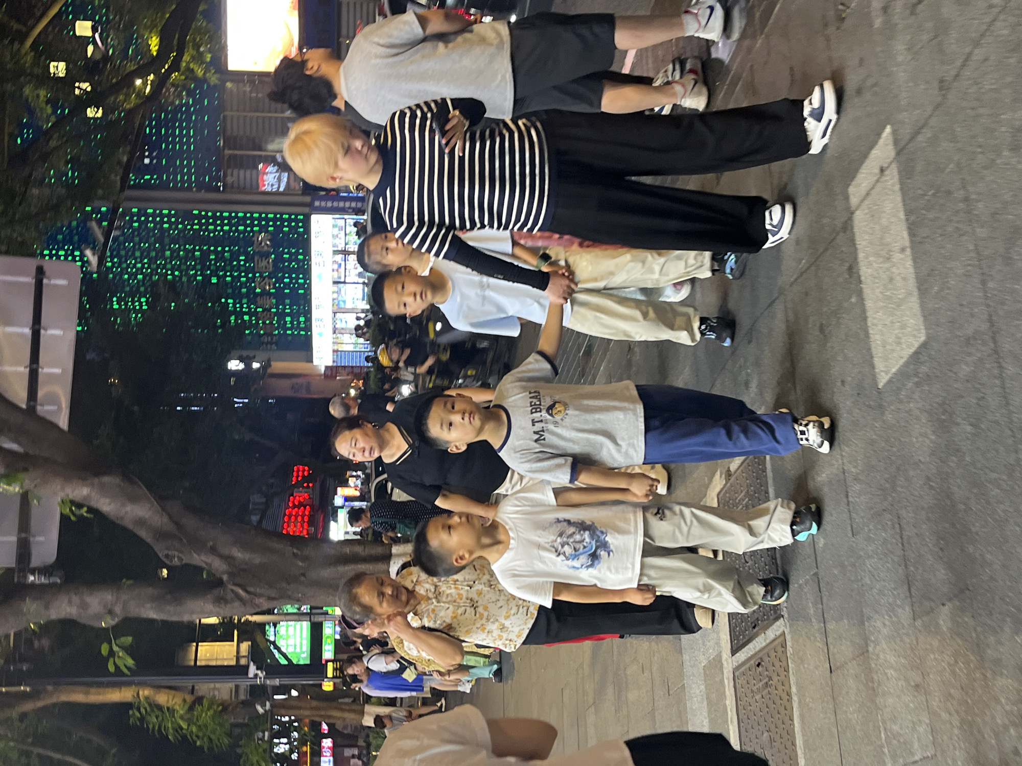 The image shows a group of people standing on a paved street at night. The scene is illuminated by streetlights and neon signs, creating a vibrant and somewhat blurry atmosphere. The people are a mix of ages, including adults and children, and they appear to be standing together, possibly as a family or group of friends.
The subjects are diverse in their attire. One person in the foreground is wearing a striped long-sleeved shirt and black pants. Another person, a child, is wearing a light-colored t-shirt with a graphic design. An older woman is wearing a patterned top. Several individuals are wearing athletic shoes.
The background features a large tree with some foliage, and behind that, buildings with illuminated windows and various signs. Some of the signs have Chinese characters on them. One prominent sign features green lights arranged in a grid pattern. Another sign displays red characters. The overall setting suggests an urban environment, likely a street or pedestrian area in a city.
The time of day is clearly night, indicated by the artificial lighting and the darkness of the sky. The weather appears to be clear, with no visible rain or fog. The people's expressions are not clearly discernible due to the lighting and resolution, but they seem to be calmly standing. There is no specific event clearly depicted, but the gathering of people suggests a social interaction.
Visible text includes "M.T.BEAF" on a child's shirt, possibly a brand or inscription. Some of the signs in the background also contain Chinese characters, but they are not legible enough to be transcribed.