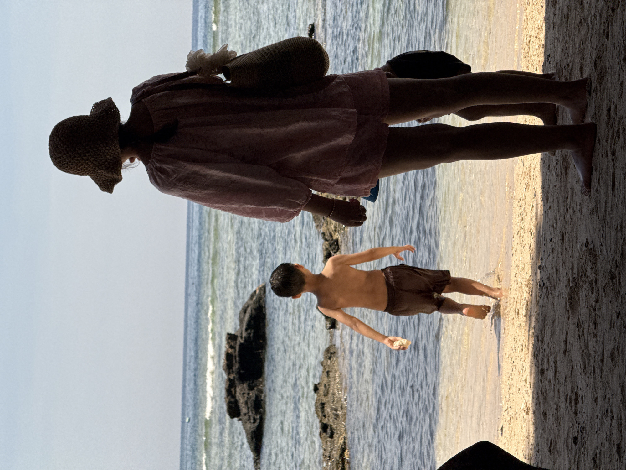 The image depicts a beach scene on Lý Sơn Island, Vietnam. In the foreground, two figures are partially visible, silhouetted against the bright background. To the right stands a person wearing a straw hat and a light pink outfit, carrying a woven bag. Their back is to the camera, and they appear to be looking out towards the ocean. To the left of this figure, a young boy is walking into the shallow water. He is wearing dark shorts and is holding a light-colored object in his right hand. The water is clear with visible waves and dark rocks jutting out of the sea in the mid-ground. The beach itself is sandy, with a gentle slope towards the water. The sky is a pale blue, indicating a clear day. The overall impression is one of a relaxed, summery day at the beach.