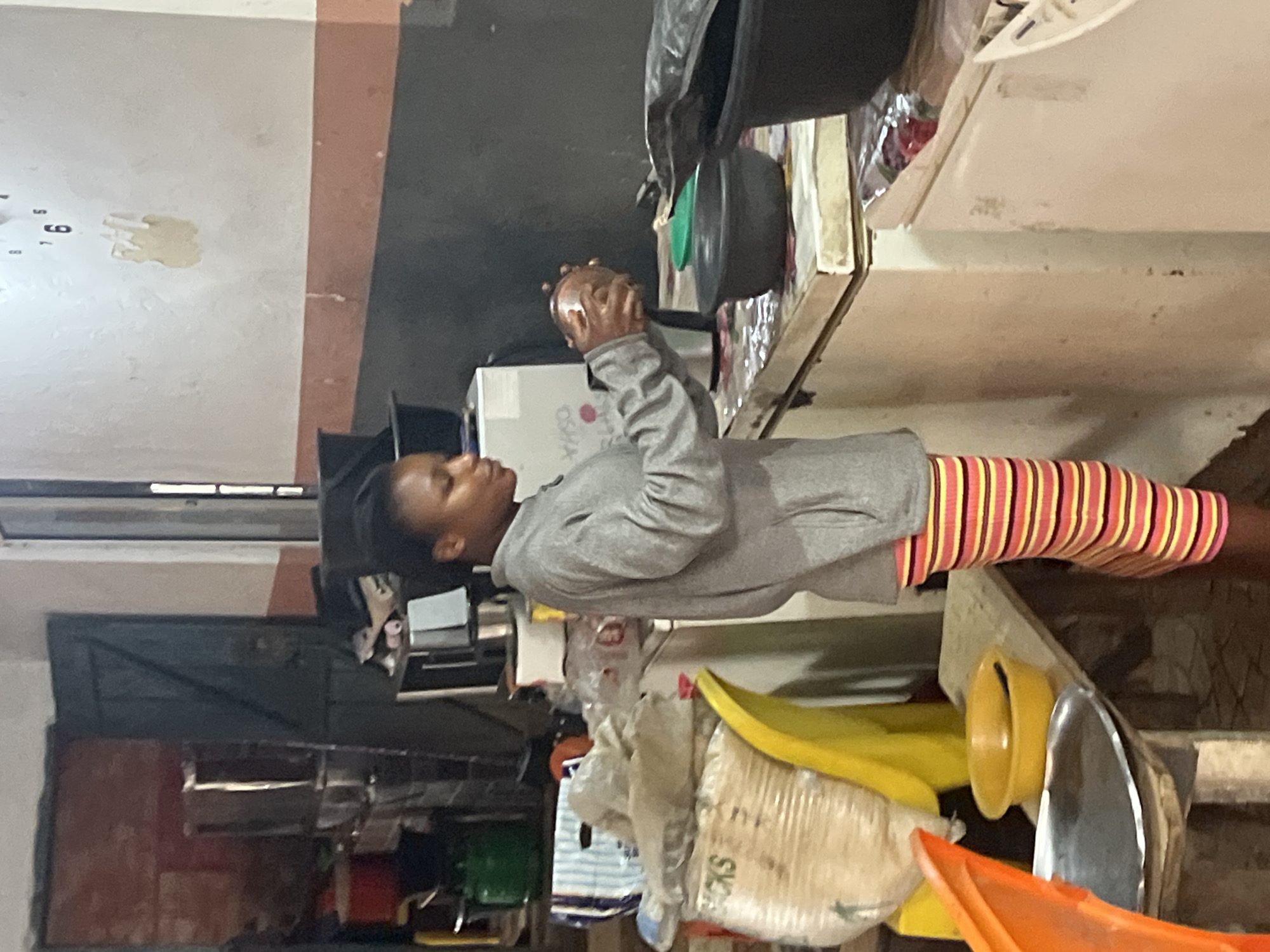 A young Black woman stands in what appears to be a market stall or food preparation area in Nkpor, Nigeria. She is wearing a grey long-sleeved jacket over a brightly striped skirt with horizontal bands of pink, yellow, and orange. Her hair is styled in a close crop. She is holding a small, round, brown object, possibly a fruit or vegetable, in her hands, and appears to be examining or preparing it.
The environment suggests a working area. To her left, a large, pale yellow, open plastic container is visible. Behind her, a large bag with visible text, including "SACKS," is leaning against a yellow surface. Further back, several shelves are laden with various items, including large containers, bags, and what seem to be food products. There are also some green and red items, potentially food or containers, on the shelves.
In front of the woman, a white appliance, possibly a freezer or refrigerator, is partially visible. On top of this appliance are several items, including a black bowl, a green lid, and the woman herself. The wall behind her has a painted stripe of reddish-brown above a darker color and a clock with Roman numerals is affixed to the upper portion of the wall, with numbers 5, 6, and 7 clearly visible. Peeling paint is evident on the wall near the clock. To the far left, a dark doorway is visible, and beyond it, red-colored objects and stacked metallic items can be seen. The overall lighting suggests an indoor setting, possibly with natural light from an opening not fully visible.
