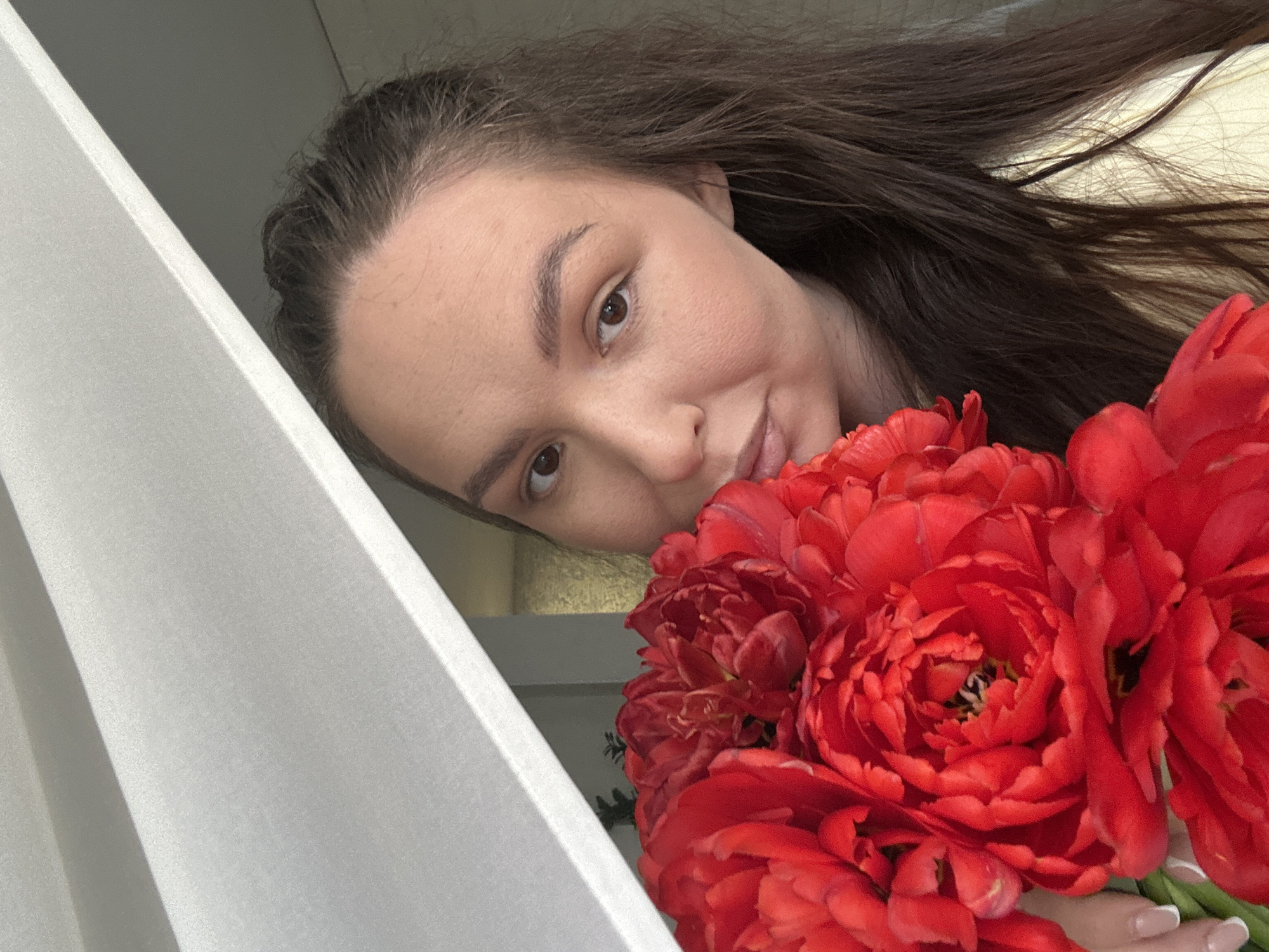 A woman with brown hair and fair skin is pictured at an angle, close to the camera. She is holding a bouquet of large, red peony-like flowers. To the left of the woman is a white, angled surface, possibly a wall or a shelf. The location is the city of Chaikovsky, Russia.