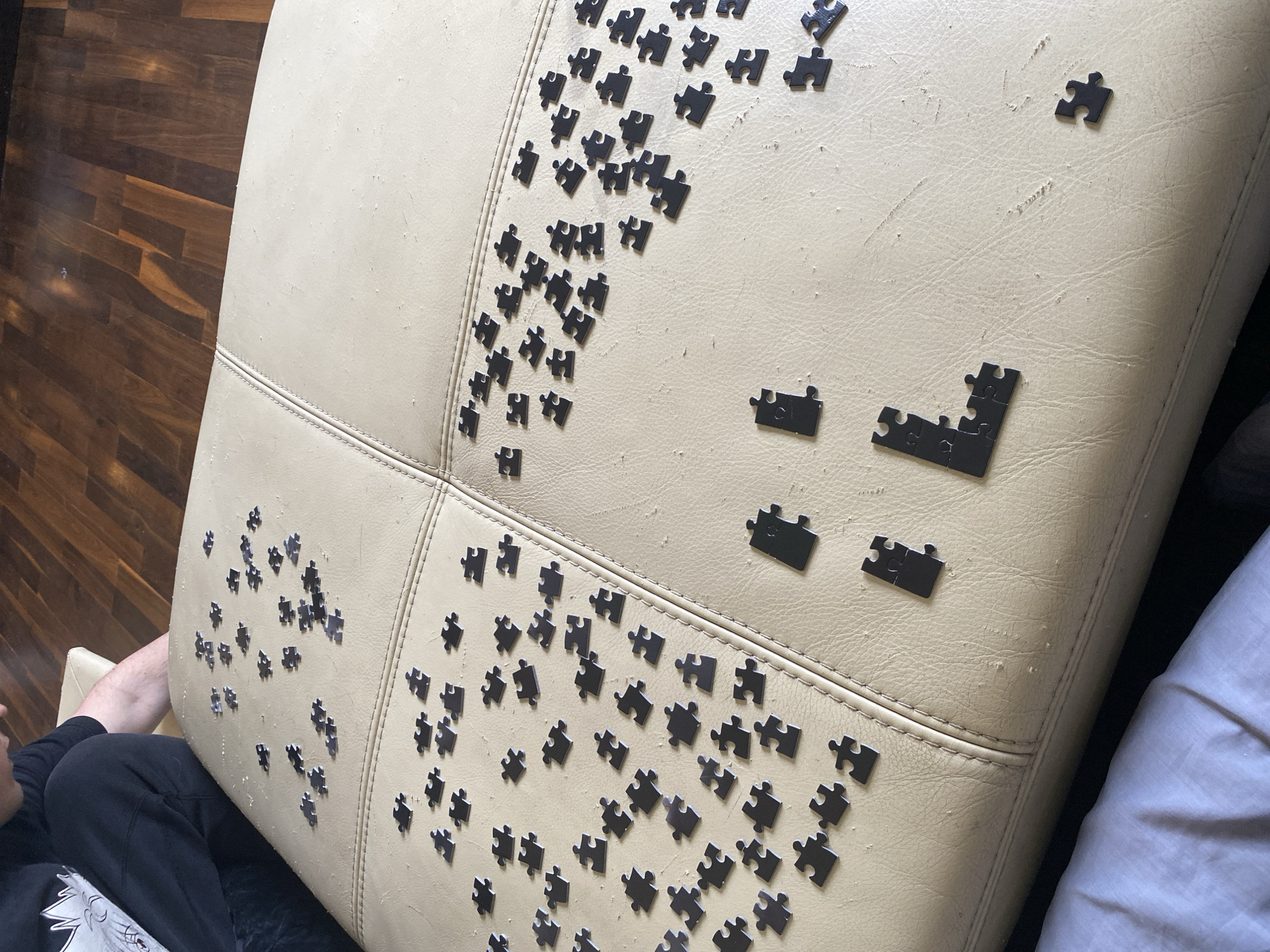 The image shows a partially assembled jigsaw puzzle laid out on a beige, leather-like surface, possibly a couch or chair. The puzzle pieces are predominantly black. A person wearing dark pants and a white graphic design on their clothing is seated in the lower-left corner of the image. The floor is visible in the upper left, featuring wooden planks. The specific location in Villaviciosa, Spain cannot be determined from the provided image.