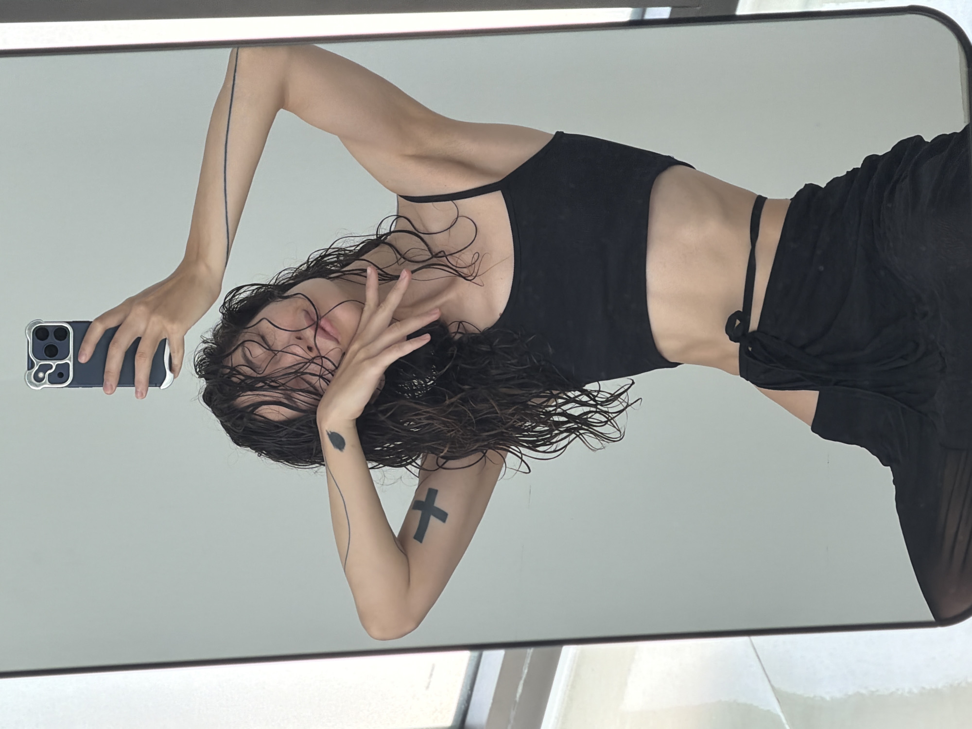 The image depicts a reflection of a person taking a selfie in a mirror. The person has wet, dark hair and is wearing a black top and bottom. They are holding a dark-colored smartphone in their left hand. The reflection shows parts of their torso and arms, revealing a cross tattoo on their left arm. There appears to be an abstract dark design on the same arm near the elbow. The background is a plain white or light gray wall. The environment suggests an indoor setting, possibly a bathroom or dressing room. There are no specific features in the reflection indicating the city of Đà Nẵng, Vietnam.