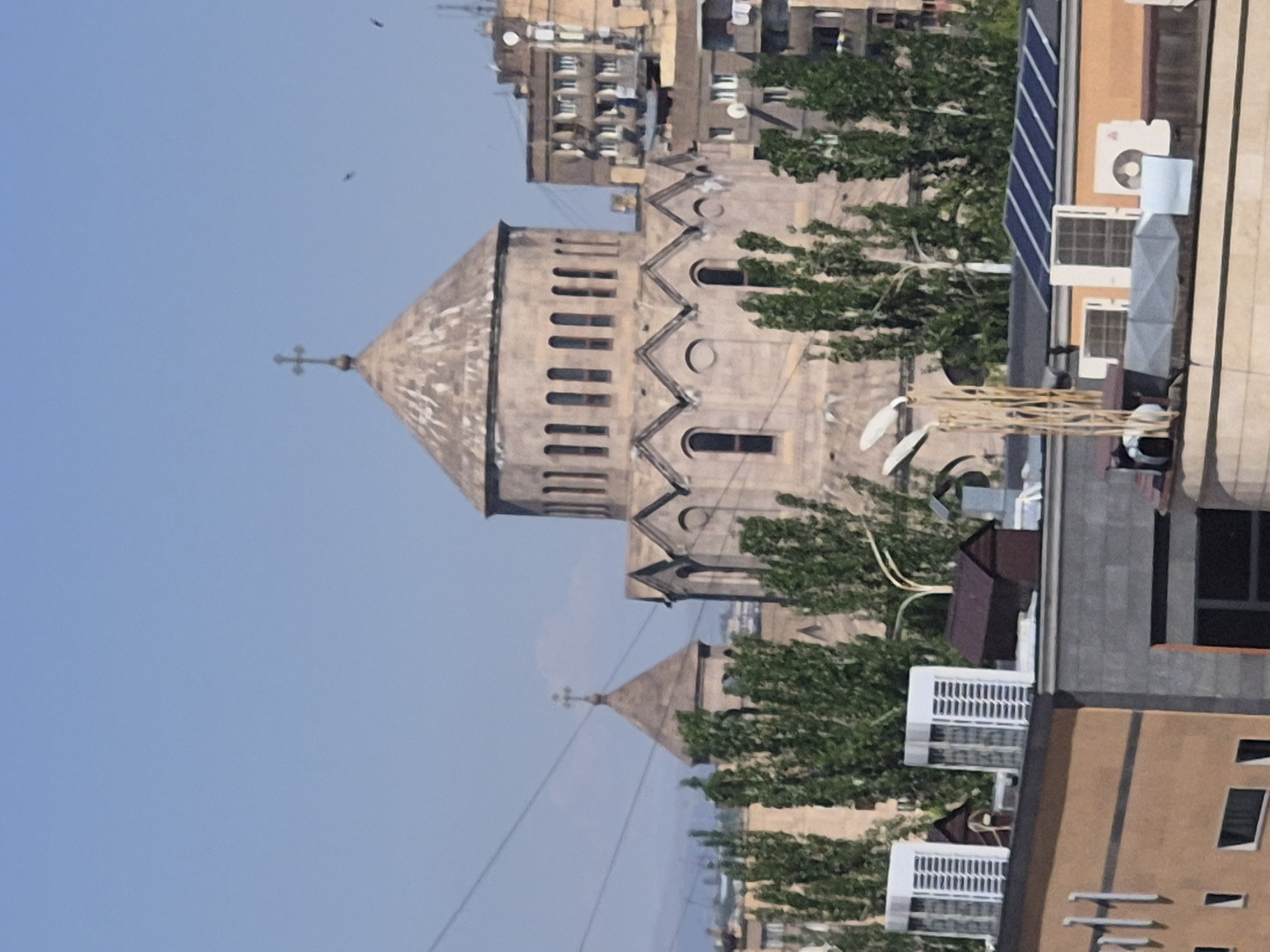 This media file captures an urban landscape in Yerevan, Armenia, featuring two prominent churches amidst residential buildings under a clear blue sky during daylight. The main subject is a large, light-colored stone church situated in the midground, characterized by a tall, octagonal drum topped with a conical dome and a cross. The church displays distinctive Armenian architecture, including a zigzag frieze and arched windows. To the left and further in the background, a smaller conical dome with a cross is visible, indicating another church.
The churches are surrounded by numerous multi-story buildings, likely residential or mixed-use, with balconies and varying architectural styles typical of a post-Soviet city. The rooftops of foreground buildings are equipped with an array of modern urban fixtures, including air conditioning units, satellite dishes, and solar panels on the building to the far right. Green trees are interspersed throughout the scene, suggesting urban greenery or small parks. The sky is bright and clear, with two small, dark specks, possibly birds or drones, visible near the main church's dome. Utility lines stretch across the lower portion of the sky. No people are visible, and there is no discernible text in the image.
