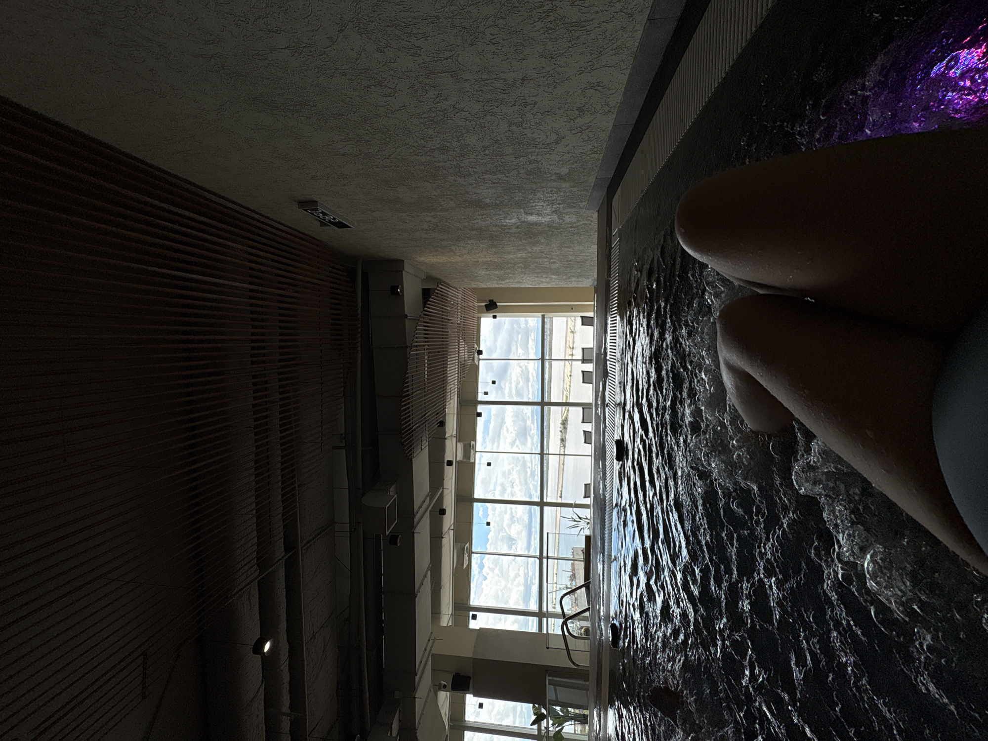 The image depicts an indoor jacuzzi with a view of a body of water and a cloudy sky through large windows. In the foreground, a person's legs are visible, submerged in the bubbly water. The jacuzzi area features a textured wall and a slatted wooden ceiling. The lighting is dim, with some purple underwater lights visible. The overall scene suggests a relaxing atmosphere. The location is identified as Khabarovsk, Russia.