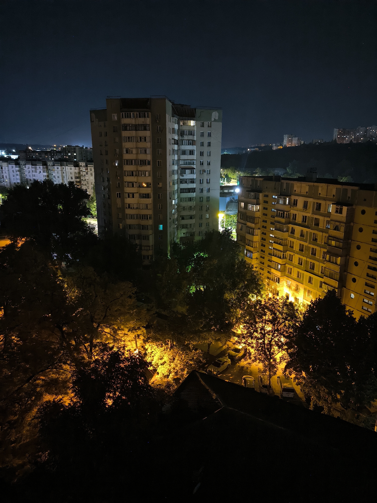 This is a nighttime, aerial view of apartment buildings and trees in Chișinău, Moldova. The scene is illuminated by artificial lights from the buildings and streetlights, casting warm, orange hues on the surrounding foliage and a parking lot with several cars. In the foreground, a dark, sloped roof is visible. The sky above is dark blue with a few faint stars. In the background, more apartment buildings and distant hills can be seen, some with lights on in the windows. The image captures a residential area at night, showcasing the contrast between the illuminated buildings and the darker natural elements.