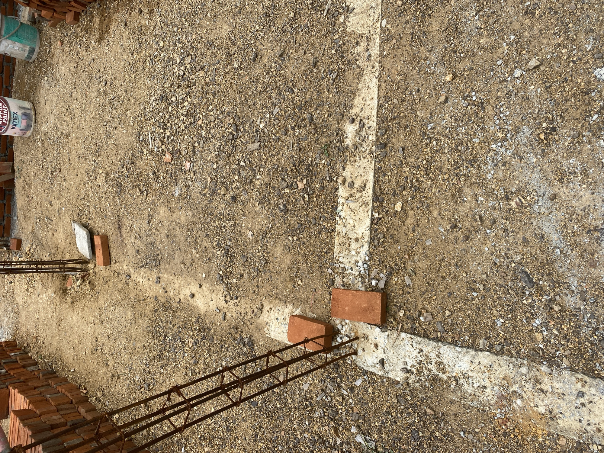 The image depicts a construction site, likely a foundation area. Piles of red bricks are visible along the left and right edges. In the center, a foundation outline is marked with white paint on the dirt and gravel ground. Rebar cages, used for reinforcing concrete, are partially embedded in the ground at intersections of the foundation lines. Two red bricks are placed on the white outline near one of the rebar structures. Two buckets, one labeled "NIPPON PAINT," are situated further back on the ground. The ground itself is a mix of soil, gravel, and small stones. The scene appears to be an outdoor location in a state of preparation for building construction. There are no people visible in the image.
