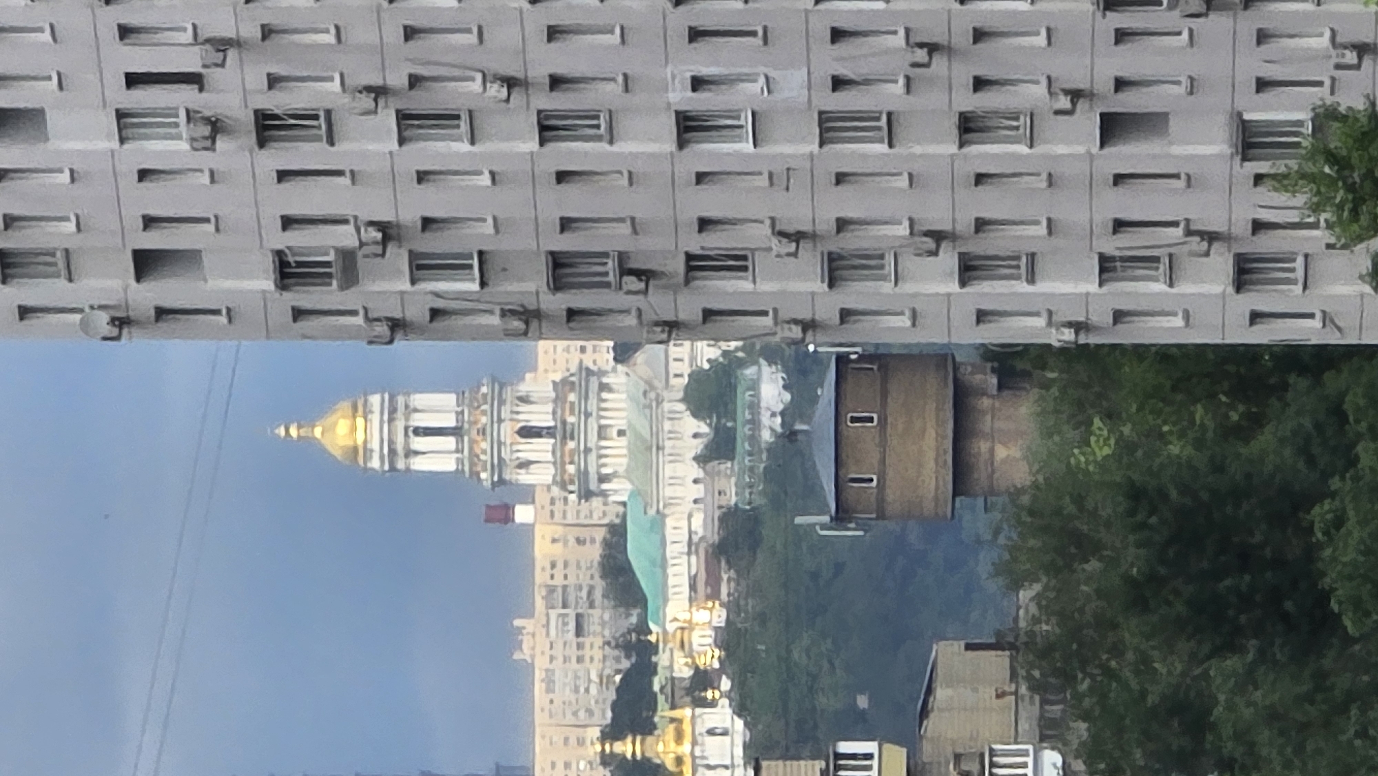 This image captures an upside-down view of an urban landscape with a distinctive architectural style. The upper portion of the image is dominated by a large, grey, multi-story building with a repeating pattern of rectangular windows. Some of these windows appear to have air conditioning units attached. Below this building, a glimpse of a golden dome atop a white, classical-style structure is visible, suggesting a church or religious building. This dome is the most prominent and brightly colored element in the scene.
Further down, a cluster of other buildings can be seen, some with a more modern, block-like appearance, while others seem to be older, possibly with decorative elements. A dense patch of green trees fills the middle ground and extends into the background, partially obscuring some of the structures. The sky is a clear, pale blue, indicating it's daytime, likely morning or late afternoon given the soft lighting. There are some dark horizontal lines in the sky, possibly power lines. The overall impression is a juxtaposition of stark, utilitarian architecture and more ornate or traditional structures, set against a natural backdrop of trees. The upside-down orientation of the image creates a surreal and disorienting effect. There is no visible text in the image.