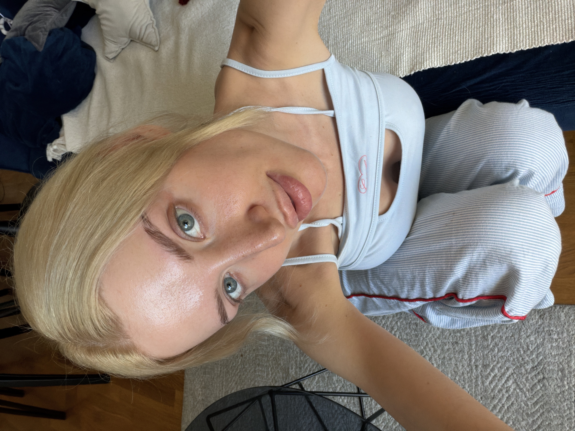A young woman with blonde hair and blue eyes is shown in a high-angle selfie. She is wearing a light blue tank top and blue and white striped pants with red trim. The background includes a beige rug with white and navy blue stripes, and a hardwood floor. A portion of a black chair is visible in the foreground. The environment appears to be an indoor domestic setting. The location in the city of Belgrade, Serbia, cannot be determined from the image.