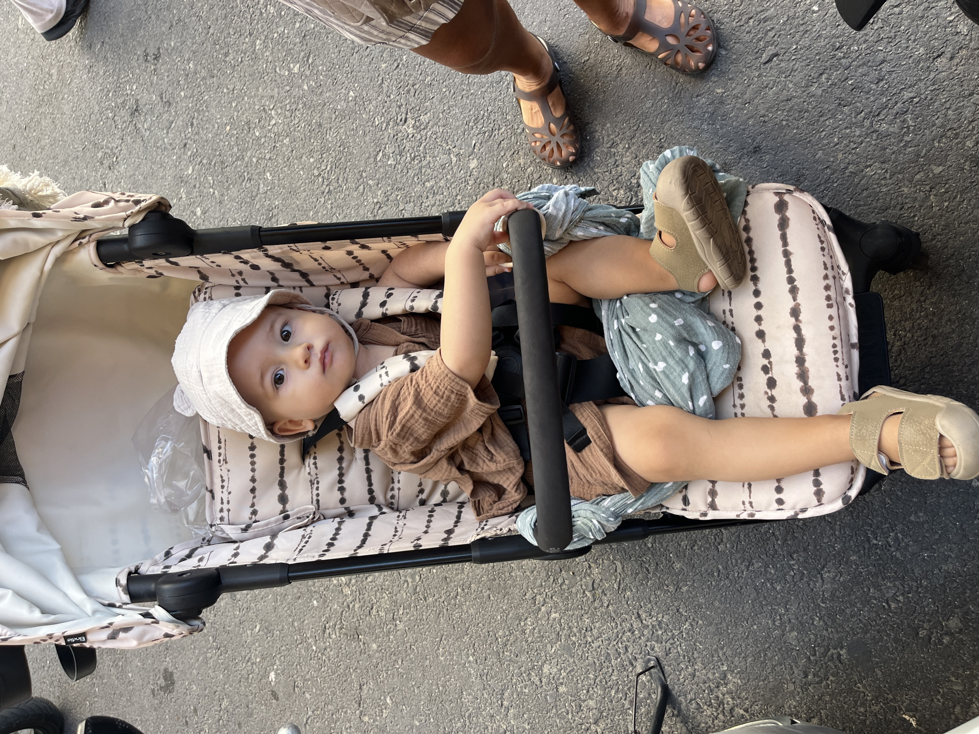 A baby lies in a stroller, oriented vertically, on an asphalt surface. The baby is wearing a tan outfit with a bucket hat and sandals. A patterned blanket is partially draped over the baby. An adult stands near the stroller. The location is Bac Nha Trang Ward, Vietnam.