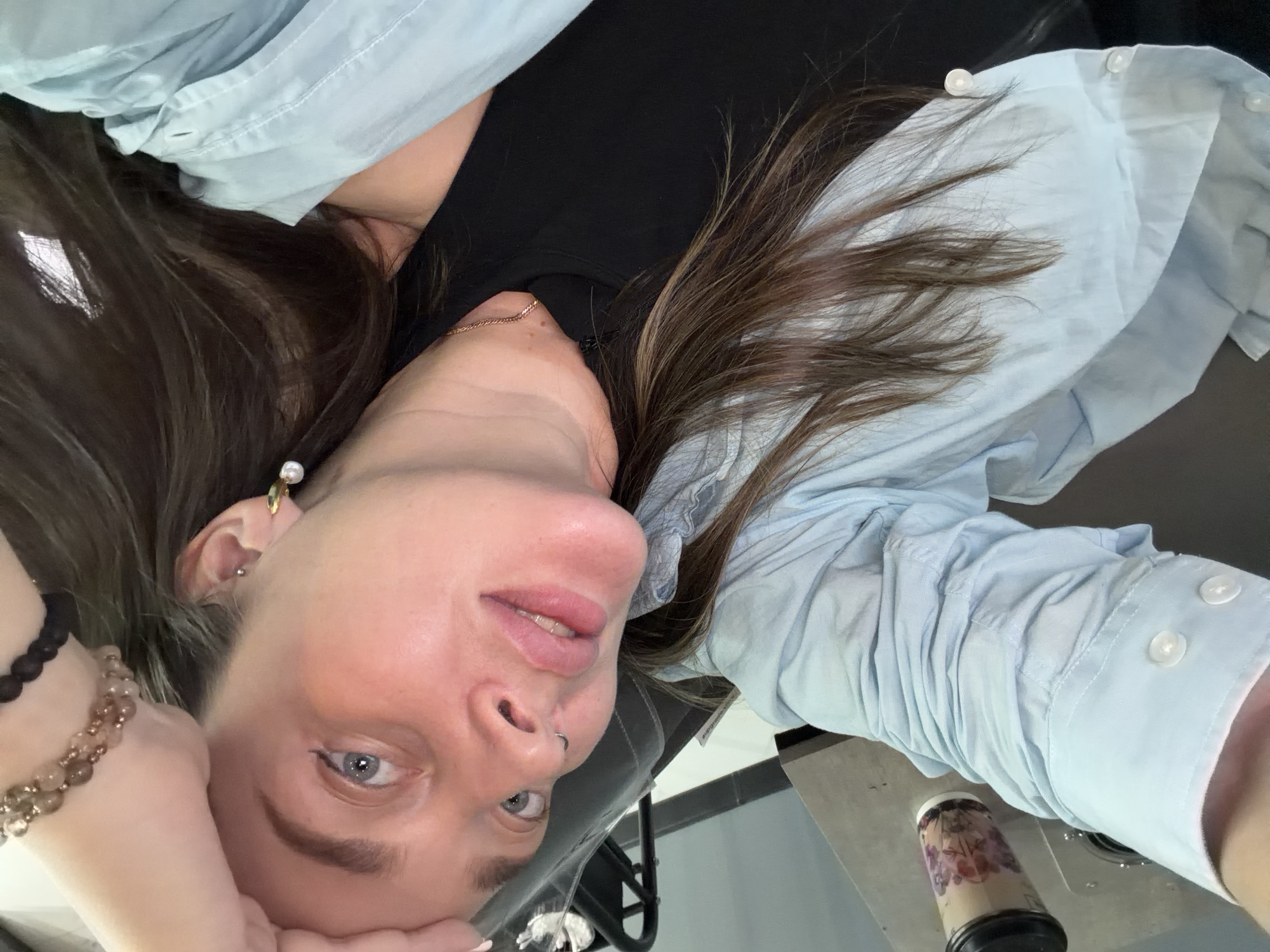 A woman with light skin and brown hair takes a selfie indoors. She wears a black top and a light blue button-down shirt. She has on a pearl earring and bracelets on her left wrist. A coffee cup sits on a silver surface to her right. The location is undeterminable.