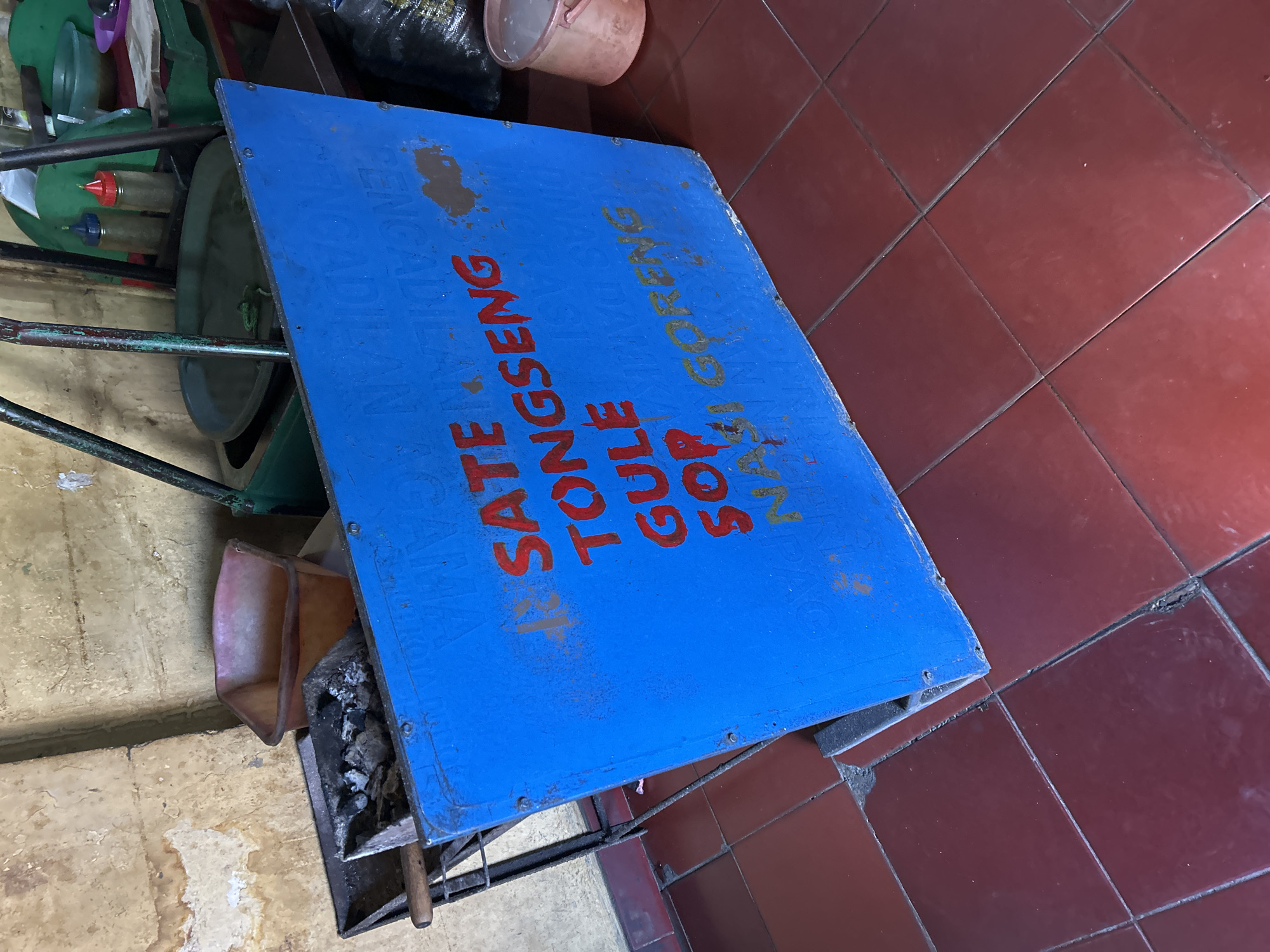 The image depicts a blue sign with red lettering advertising food items, likely at an outdoor food stall or small eatery. The text on the sign reads "SATE," "TONGSENG," "GULE," "SOP," and "NASI GORENG," which are popular Indonesian dishes. The sign is placed on a red tiled floor, typical of outdoor markets or food vendors in Indonesia. In the background, there are various cooking implements and containers, including a charcoal grill, a large pan, and bottles of condiments, suggesting active food preparation. The walls are a distressed, textured material, possibly concrete or plaster, common in older buildings or informal commercial spaces. The overall environment suggests a humble, possibly urban, setting in Depok, Indonesia, where such food stalls are common.