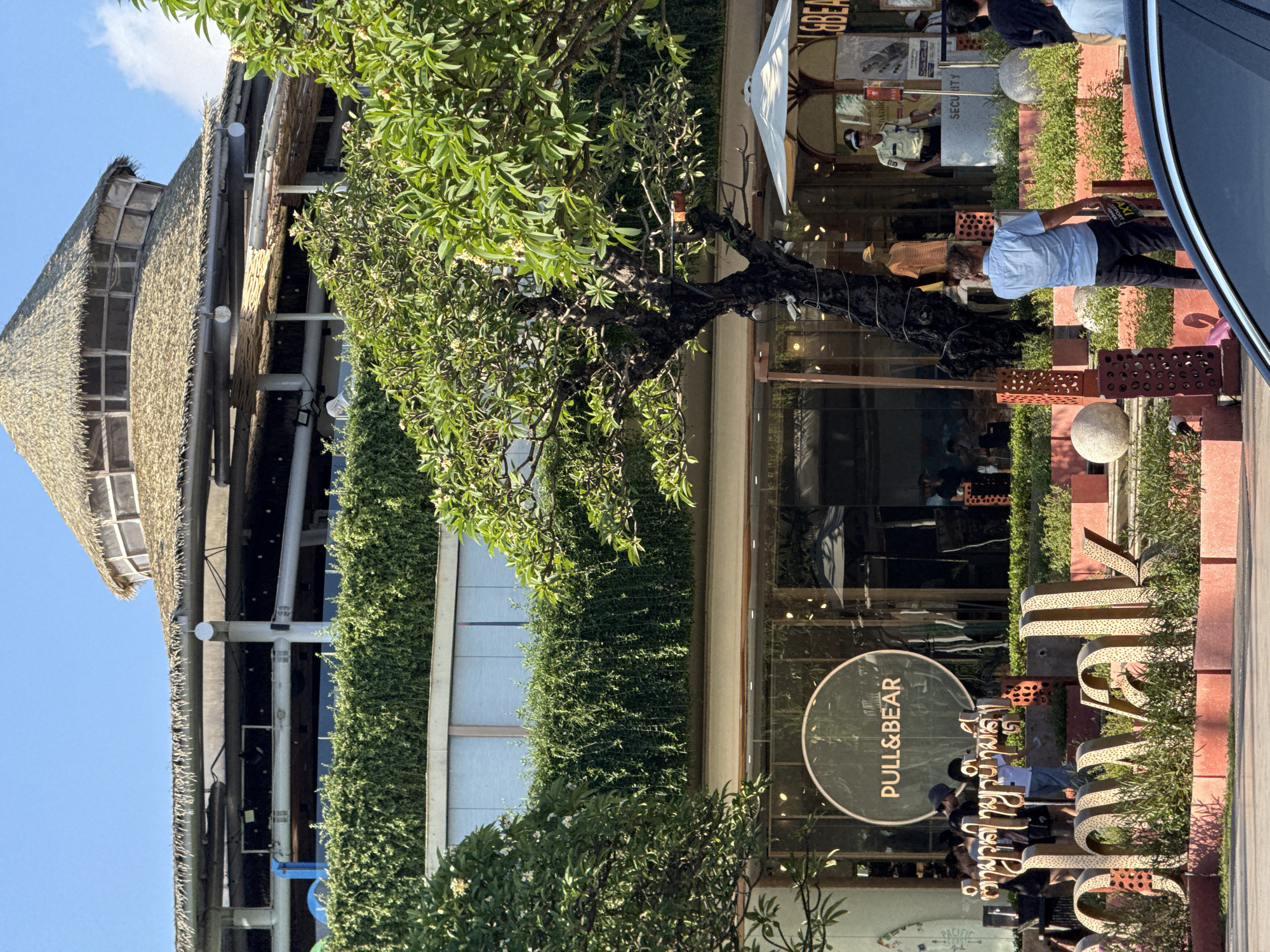 The image depicts the storefront of a Pull&Bear store located in Kuta, Indonesia. The building has a unique design, incorporating a thatched roof and vertical garden elements. The Pull&Bear logo is prominently displayed on the storefront. A tree with lush green foliage is situated in front of the store. Several people are visible, including customers and a security guard. The area around the store is paved with red bricks and decorated with plants. A car is partially visible on the right side of the image. The sky is clear blue, suggesting a sunny day.