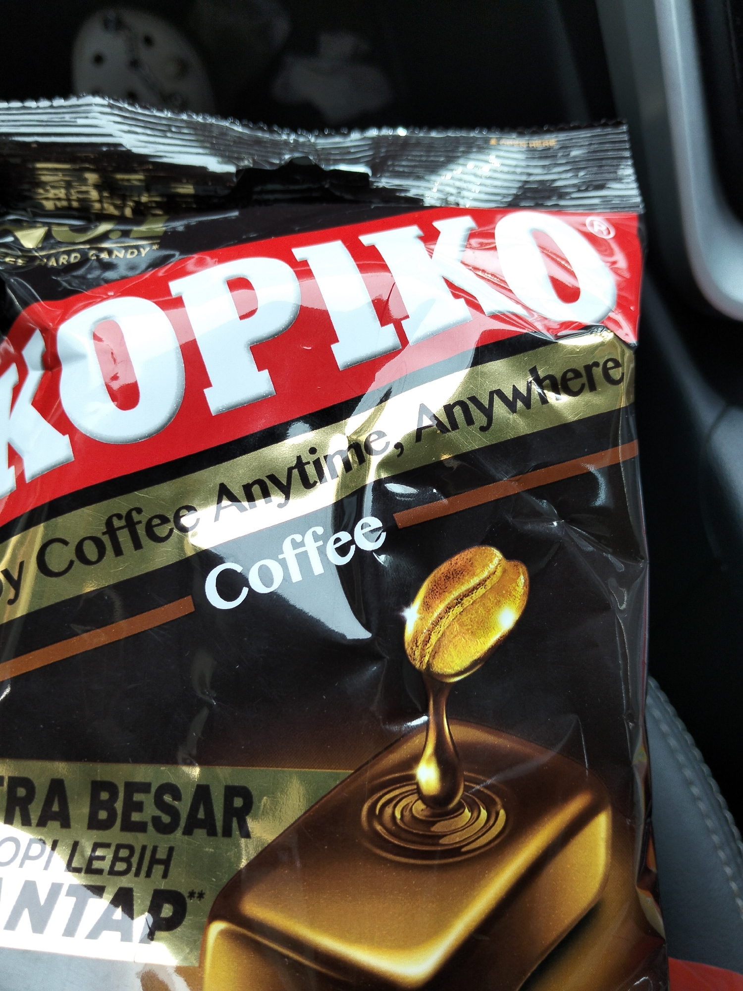 This is a close-up, eye-level, outdoor photograph of a package of Kopiko coffee candy. The package is mostly black with red and gold accents and features a large white "KOPIKO" logo. Text on the package reads "y Coffee Anytime, Anywhere" and "Coffee". Below this, there is text in a larger font that appears to be in a foreign language, followed by "NTAP**". The package is slightly crinkled and reflects light. A golden coffee bean is depicted dripping into a chocolate-like substance on the lower right side of the image. The background is dark and out of focus, suggesting the photo was taken in a dimly lit environment, possibly a car interior, as indicated by the dark, textured material on the right side of the frame. The product is in the city of Keudematangglumpangdua, Indonesia.