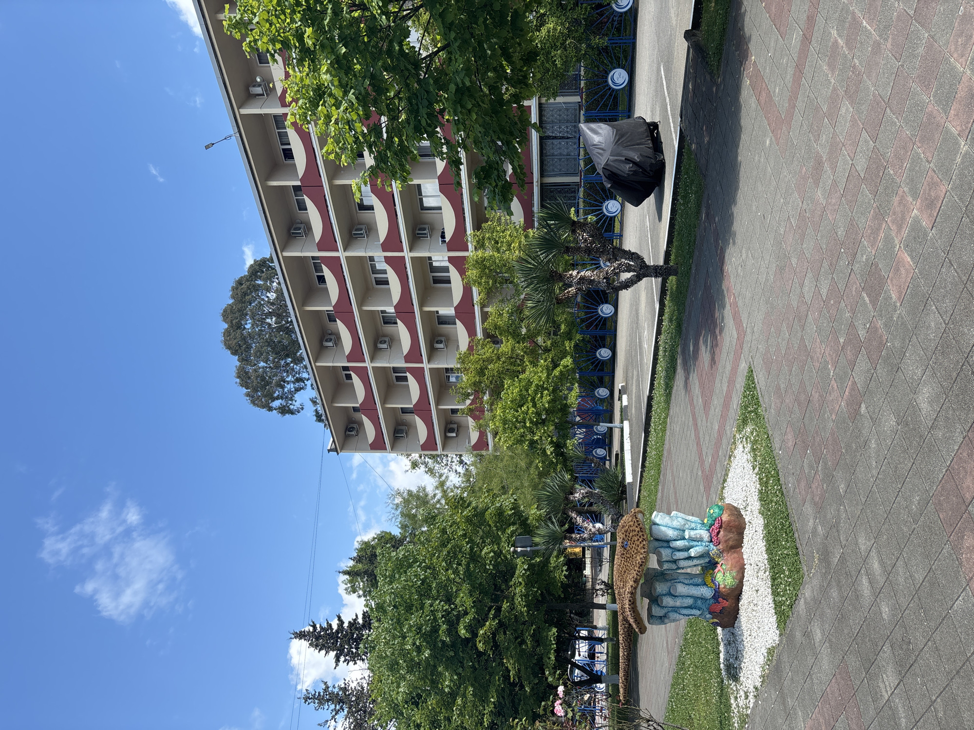 A wide shot captures a building with several floors and balconies, a paved walkway, and greenery. The building, a hotel in Sochi, Russia, features a light-colored facade with repeating red and beige balcony railings. Rows of air conditioning units are visible on the building's exterior.
To the left of the building, a small, colorful sculpture resembling a mushroom or coral formation stands on a patch of white pebbles and grass. A paved walkway, made of gray and reddish-brown interlocking bricks, stretches across the foreground. Beyond the walkway, a blue fence with a circular wave-like design separates the area from a parking lot. In the parking lot, a motorcycle covered with a black tarp is parked. Various trees, including palm trees, are scattered throughout the scene, adding to the lush, green environment. The sky above is bright blue with scattered white clouds. The overall scene is peaceful and appears to be a sunny day.