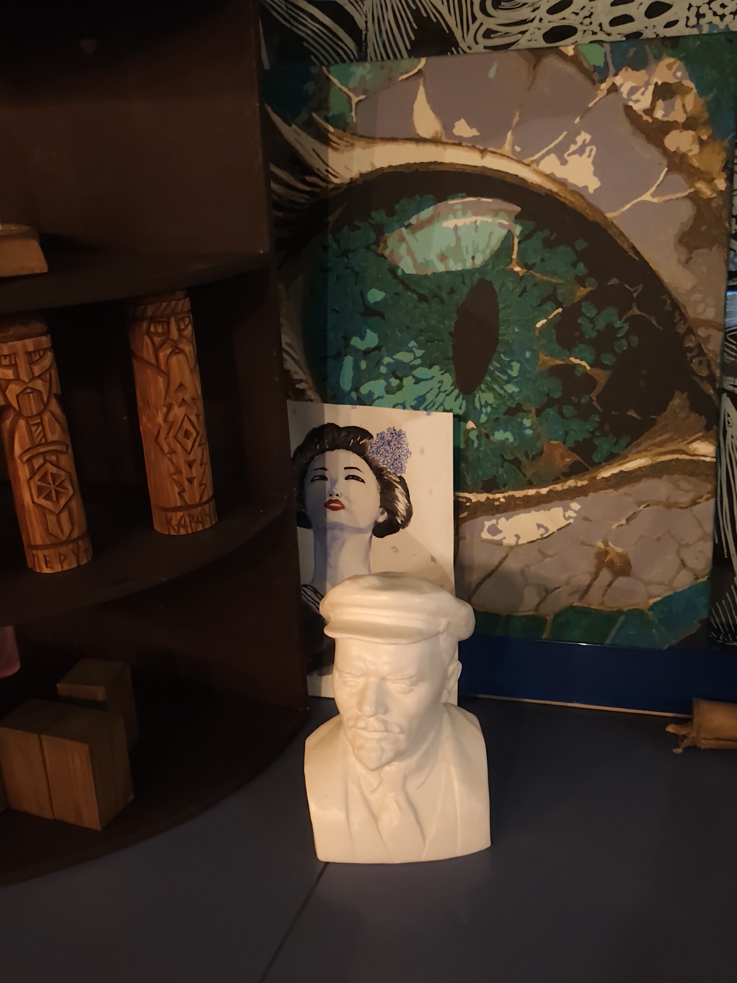 The image depicts an indoor scene, possibly a room or a display area, featuring several decorative objects. In the foreground, a white bust of a man wearing a hat and suit is placed on a dark blue surface. Behind the bust, to the left, are three carved wooden figures on a dark shelf. To the right of the bust, a framed artwork, possibly a painting or print, is leaning against a surface. The artwork appears to be a close-up of an eye with intricate patterns. Further to the left, another artwork featuring a portrait of a woman with dark hair and a flower in her hair is visible. Wooden blocks are also present on the shelf to the left. The overall impression is of a collection of art and decorative items. The image does not provide any specific details about the city of Sochi, Russia, or any actions or interactions between people.