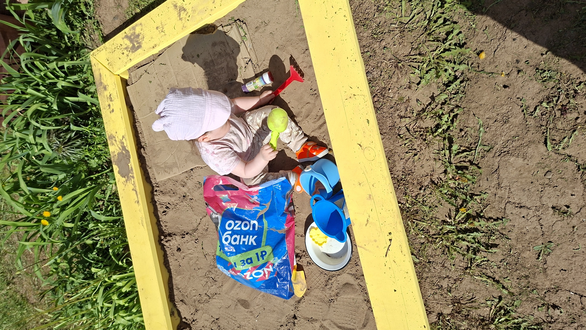 Observed in Большой Букор, Russia, is a child playing in a yellow-framed sandbox. The child, wearing a light-colored head covering and clothing, sits amidst various toys including a green scoop, a red rake, and blue buckets. A white dish and a plastic bag with Russian text "ozon банк" are also within the sandbox. The sandbox sits on soil interspersed with grass and weeds. Shadows indicate the presence of sunlight.