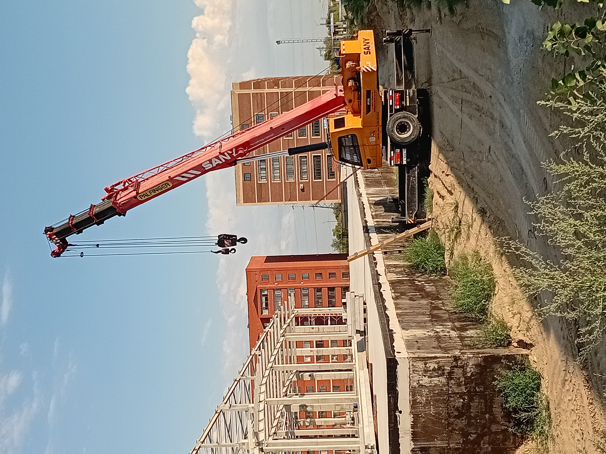 A SANY brand telescopic mobile crane, colored orange and red, is positioned on a dirt construction site. The crane's boom is extended upwards and to the right. In the background, there are two multi-story brick apartment buildings, one red and one lighter brick color. To the left, a white metal framework for a new building under construction is visible. The sky is blue with scattered white clouds. The ground is a mix of dirt, sand, and some green vegetation. The scene appears to be a construction project in the city of Semey, Kazakhstan. There are no people visible in the image.