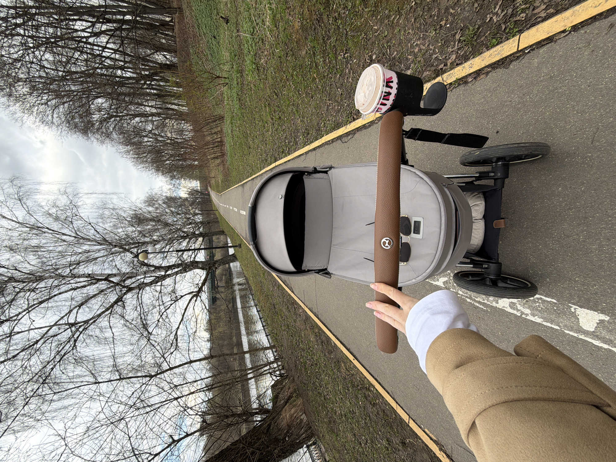 From a first-person perspective, the image captures a person pushing a baby stroller along an asphalt path in what appears to be a park or riverside area in Khimki, Russia. The path, marked with a continuous yellow line on the right and a broken white line in the middle, stretches into the distance.
The person's hand, wearing a white sleeve and a light brown coat, holds the brown faux-leather handle of a grey baby stroller. A black cup holder is attached to the stroller handle, holding a paper coffee cup with a white lid. Visible text on the cup reads "VANA" in red lettering. A silver circular logo with an intertwined symbol, resembling the Cybex brand, is prominent on the handle. Additionally, a black tag on the stroller's fabric reveals the text "RECARO S-LINE".
The immediate surroundings show sparse green and brown ground cover beside the path. Both sides of the path are lined with numerous bare-branched trees, suggesting late winter or early spring. Through the trees on the left, a body of water, likely a river or large pond, is visible under an overcast sky. The overall scene conveys a calm, leisurely outdoor stroll on a cloudy day.