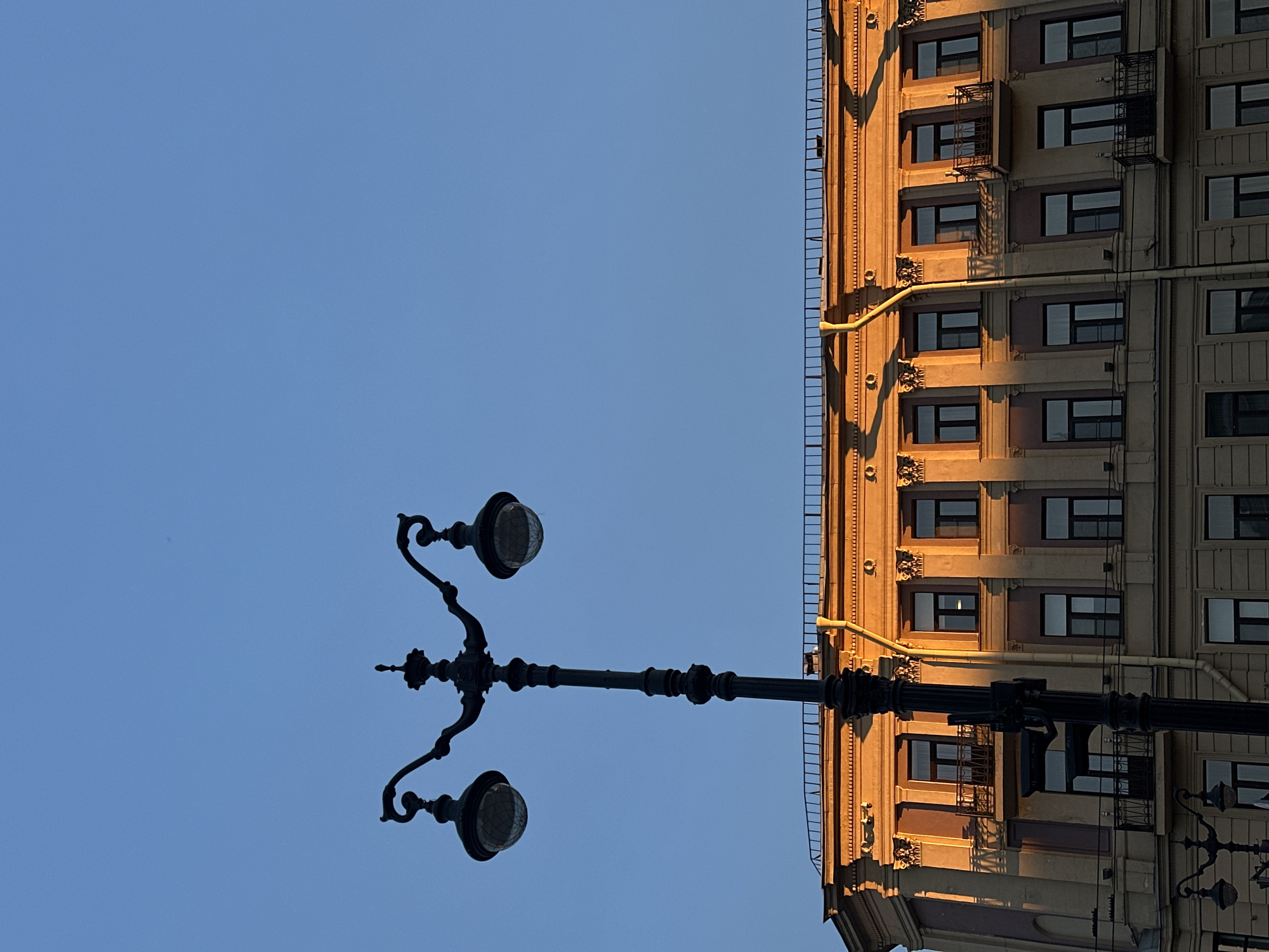 The image shows a low-angle view of a vintage lamppost against a clear blue sky, positioned next to a multi-story building facade. The building's architectural style features repetitive window patterns, ornate detailing, and vertical piping. Sunlight casts shadows across the building's surface, highlighting its texture. The lamppost is black and has two ornate lamps. The orientation of the image is rotated.