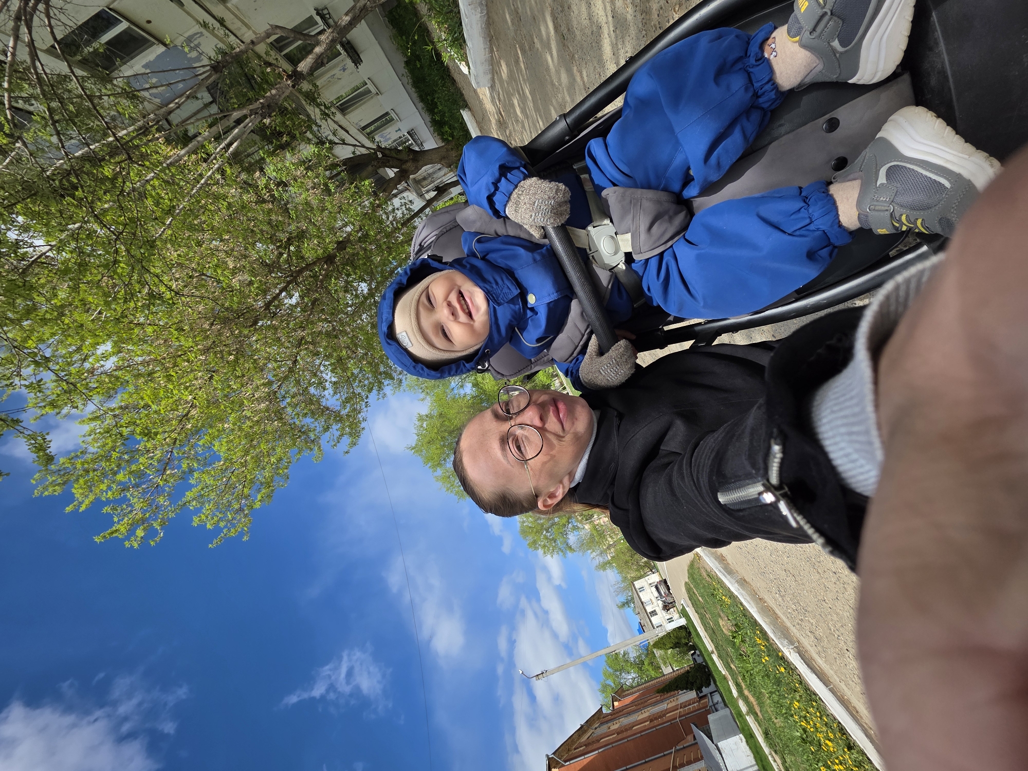 A father takes a selfie with his child in a stroller. The location appears to be outdoors in a city setting. The child is wearing a blue jumpsuit with mittens and gray sneakers. The father is wearing a black jacket and glasses. Buildings and trees are visible in the background. The sky is blue with some clouds. The location is plausibly Chaikovsky, Russia, given the architecture.