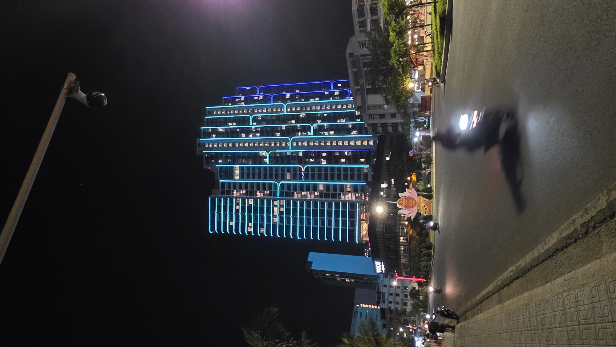 A night-time view of Bắc Nha Trang, Vietnam. The main subject is a tall, modern building illuminated with blue neon lights highlighting its architectural features. The building is multi-storied and has many windows, some of which are illuminated from within. A street runs in front of the building, with a blurred motorcycle speeding along it. Other buildings and structures can be seen in the background. A camera is visible attached to a pole on the left of the image. The overall environment is urban, characterized by the contrast of the brightly lit buildings against the dark night sky.