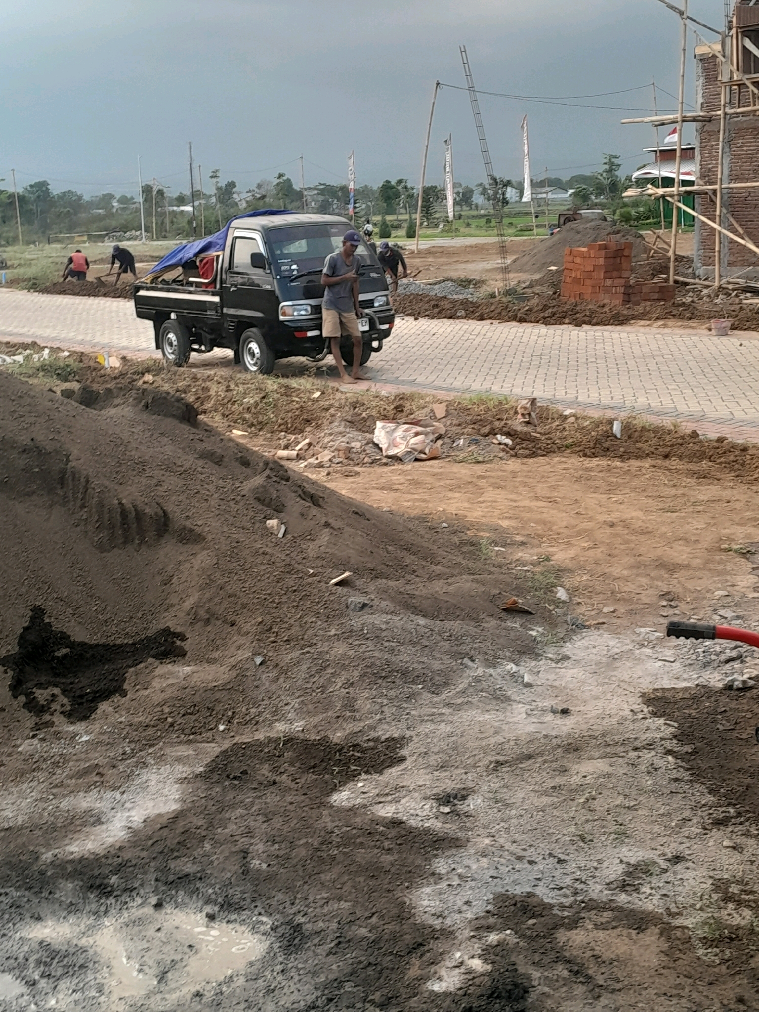 A black pickup truck is parked on a paved surface next to a construction site in Karangduren, Indonesia. A man wearing a blue cap and tan shorts stands next to the truck, looking at his phone. In the background, several other individuals are working on the site, some digging and others near piles of dirt and bricks. Construction materials and scaffolding are visible, indicating ongoing building activity. The environment appears to be an outdoor area with a cloudy sky and some vegetation.